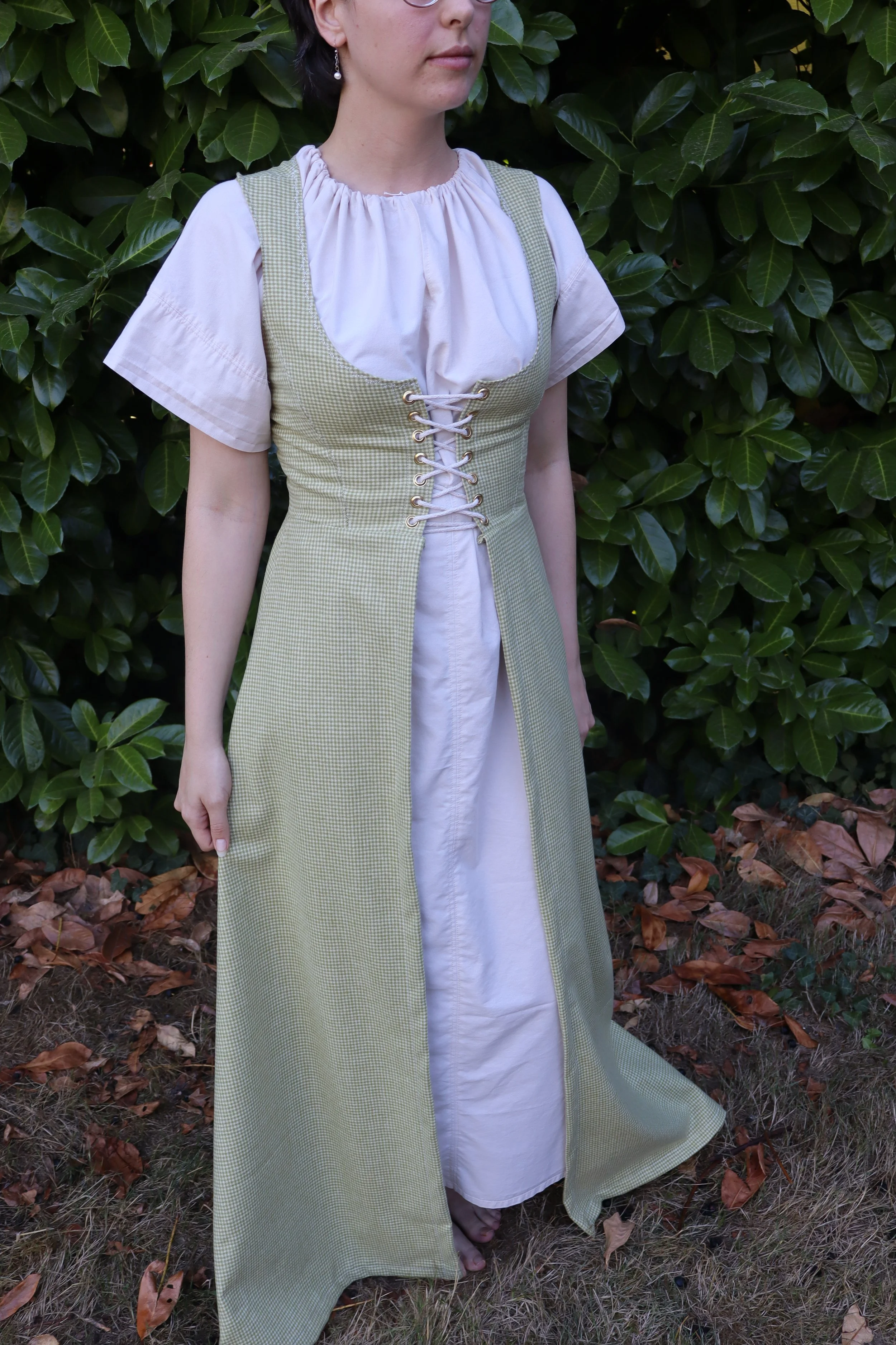 woman with glasses wearing a green overdress standing in front of green foliage. Garment is medieval inspired, historically influenced renaissance faire clothing.