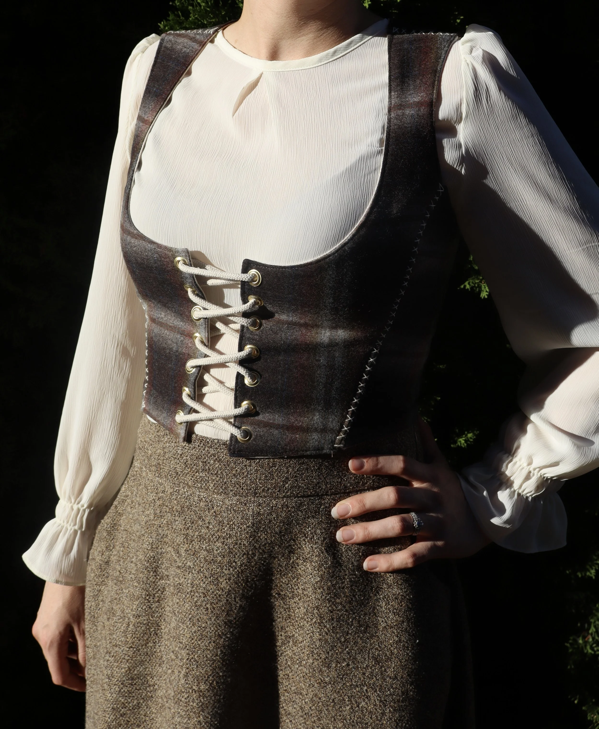 A woman wearing a beige blouse, plaid vest, and brown high-waisted skirt with her hand on her hip.