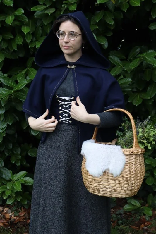 A woman with glasses and dark hair standing outdoors in front of green foliage, wearing a dark hooded cape made of 100% wool, a gray dress with black lacing, and holding a wicker basket with a white cloth and greenery inside.