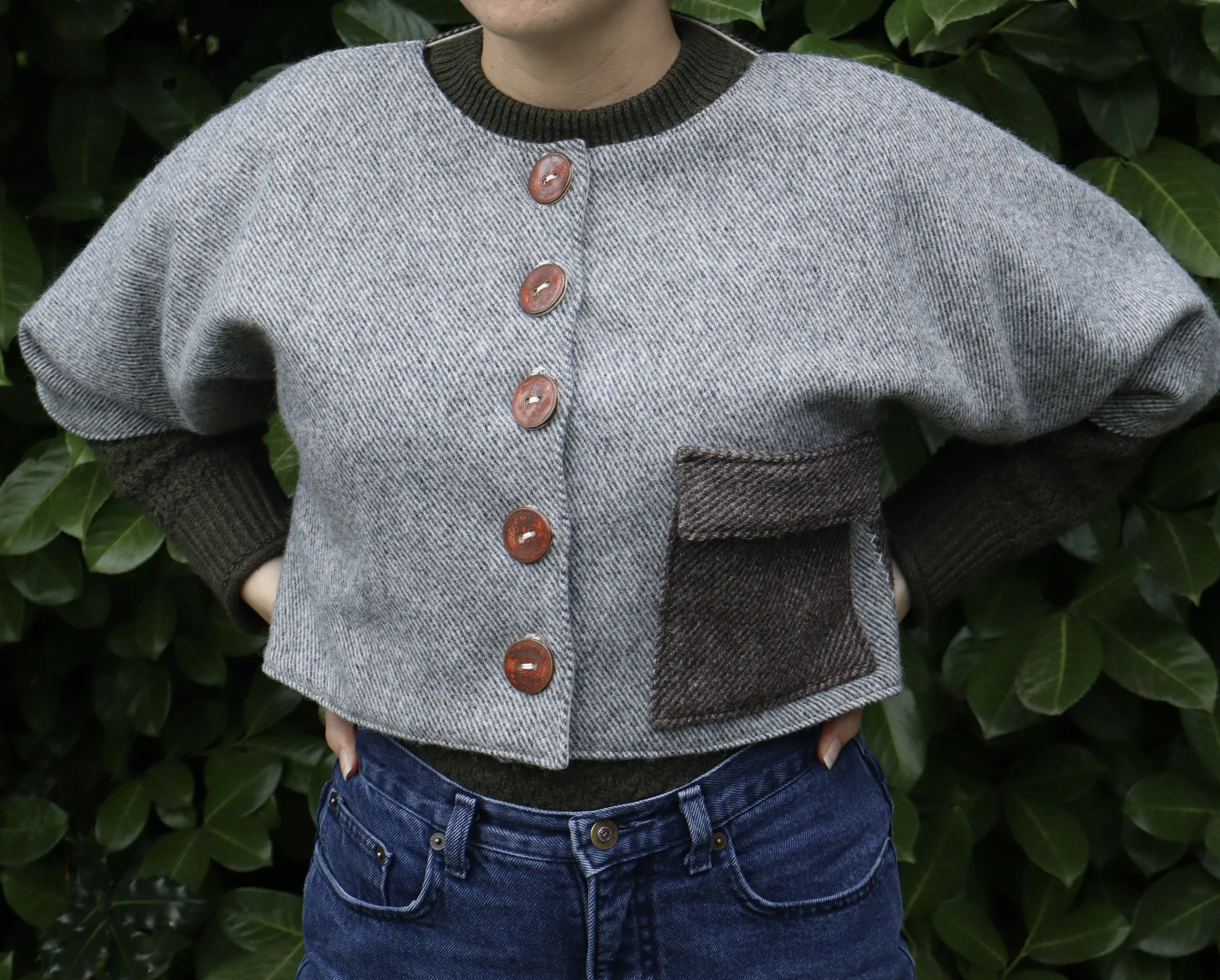 Woman wearing a light grey coat / jacket with brown buttons standing in front of green foliage. Item is medieval inspired and historically influenced