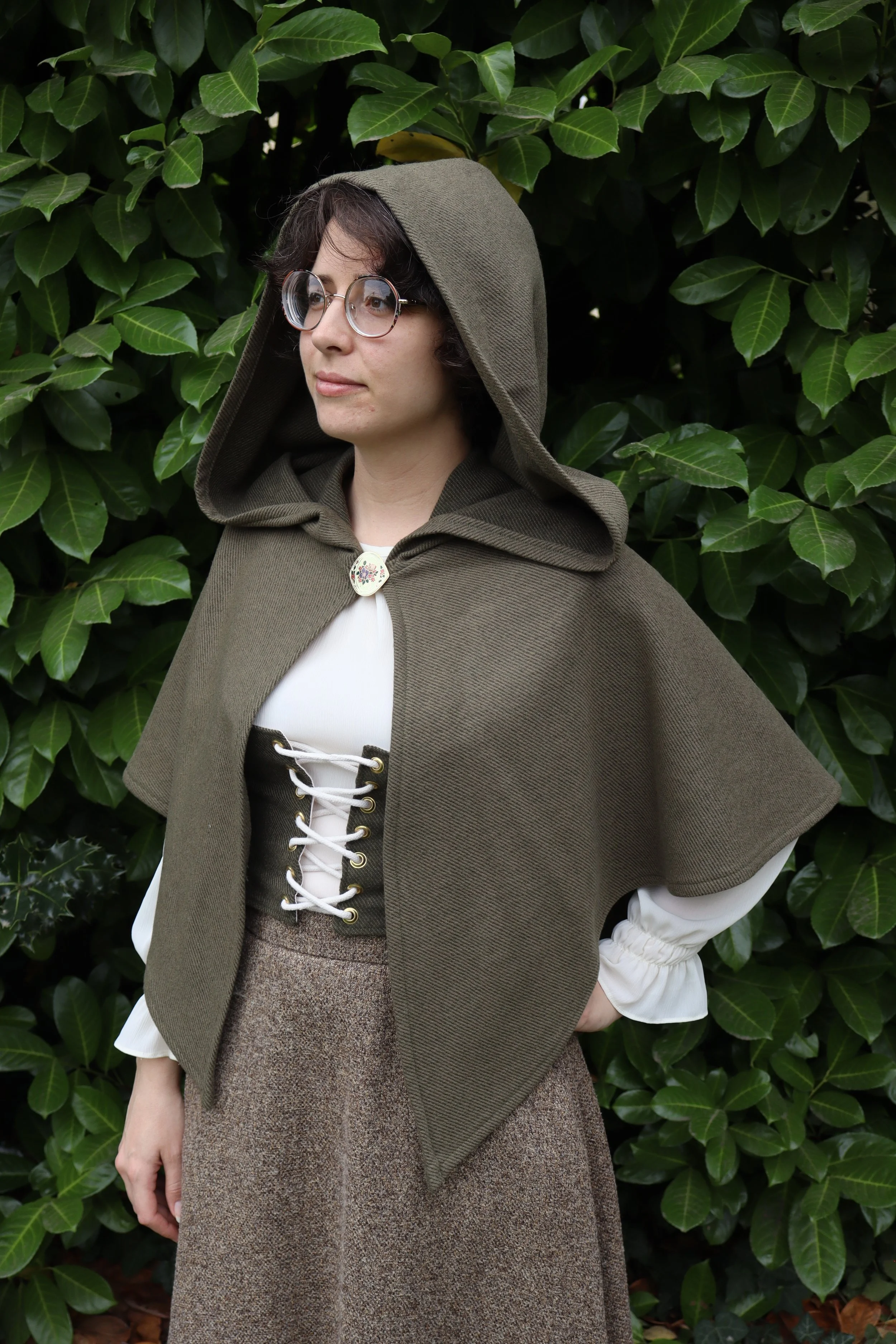 woman with glasses in a dark green capelet/cloak  and a white shirt standing in front of green foliage.