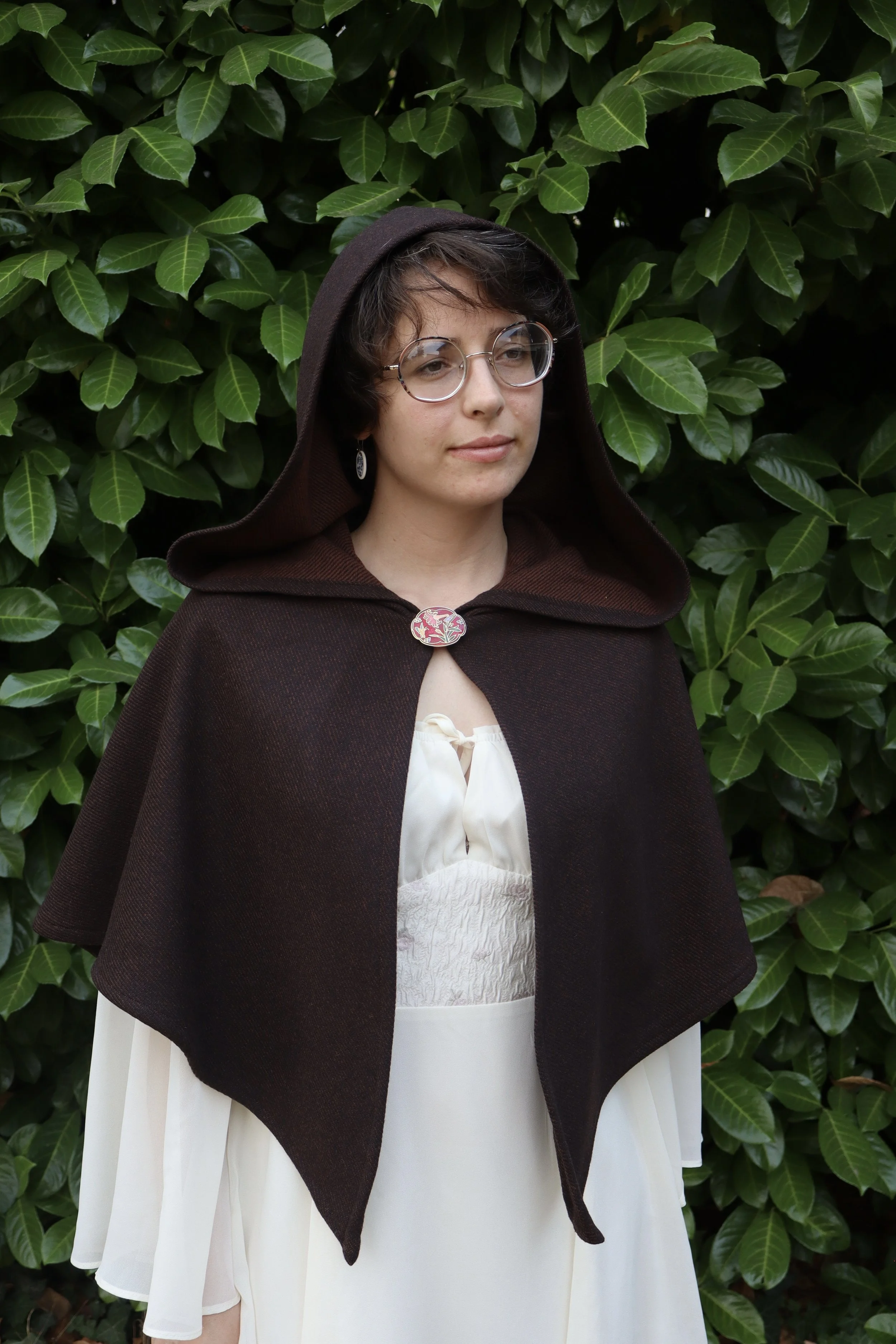 woman with glasses in a brown capelet/cloak  and a white shirt standing in front of green foliage.