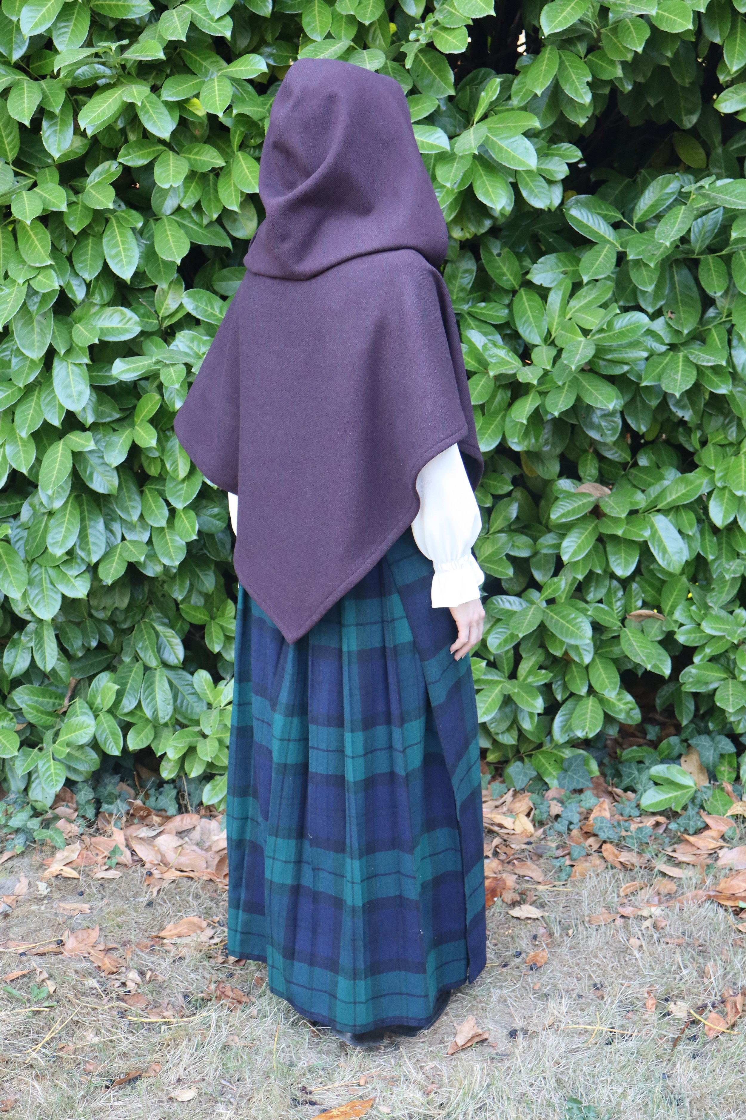 woman in a dark purple capelet/cloak facing away from the camera and standing in front of green foliage.