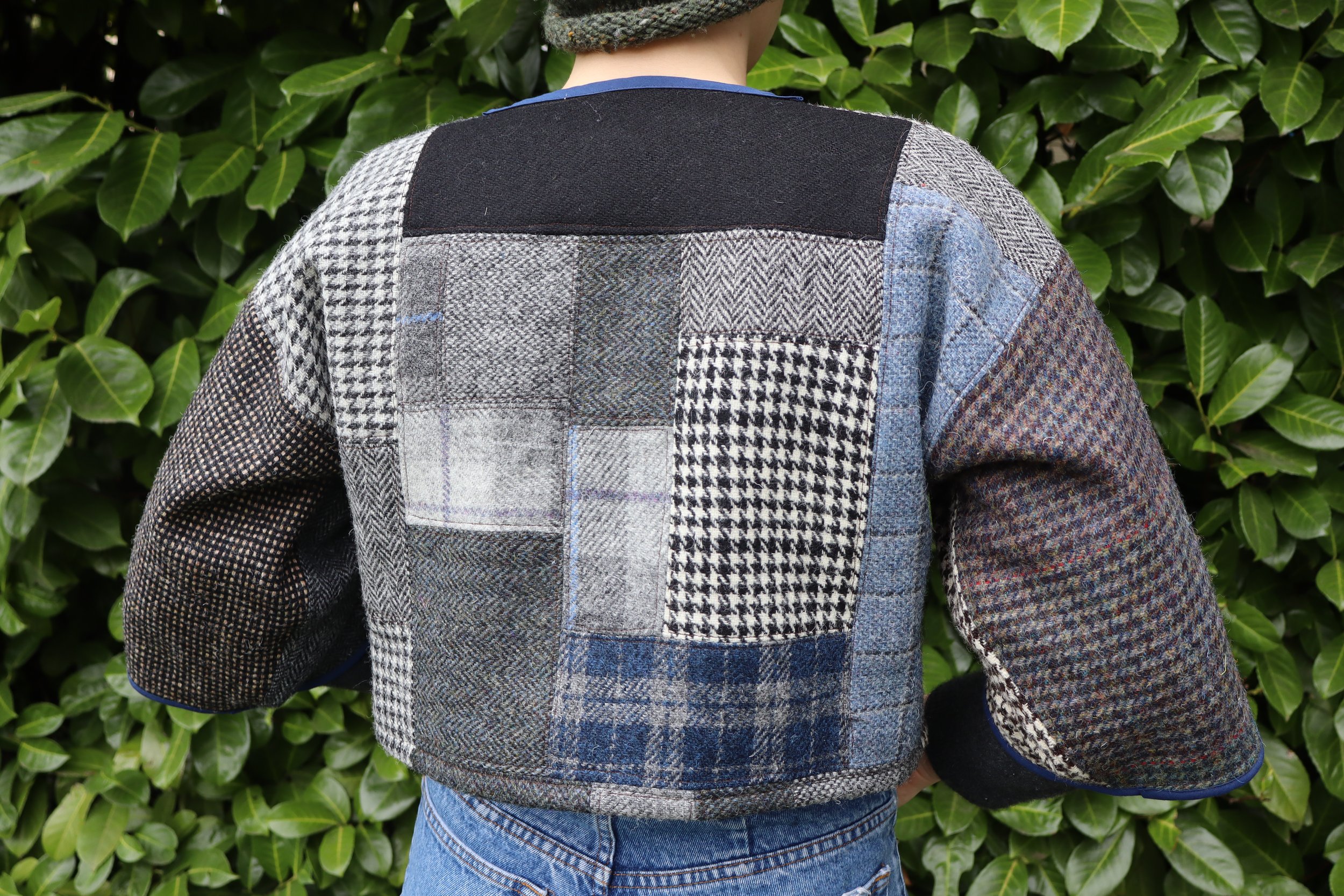 Person wearing a patchwork gray and black quilted jacket with various plaid and houndstooth patterns, standing in front of green leafy bushes.