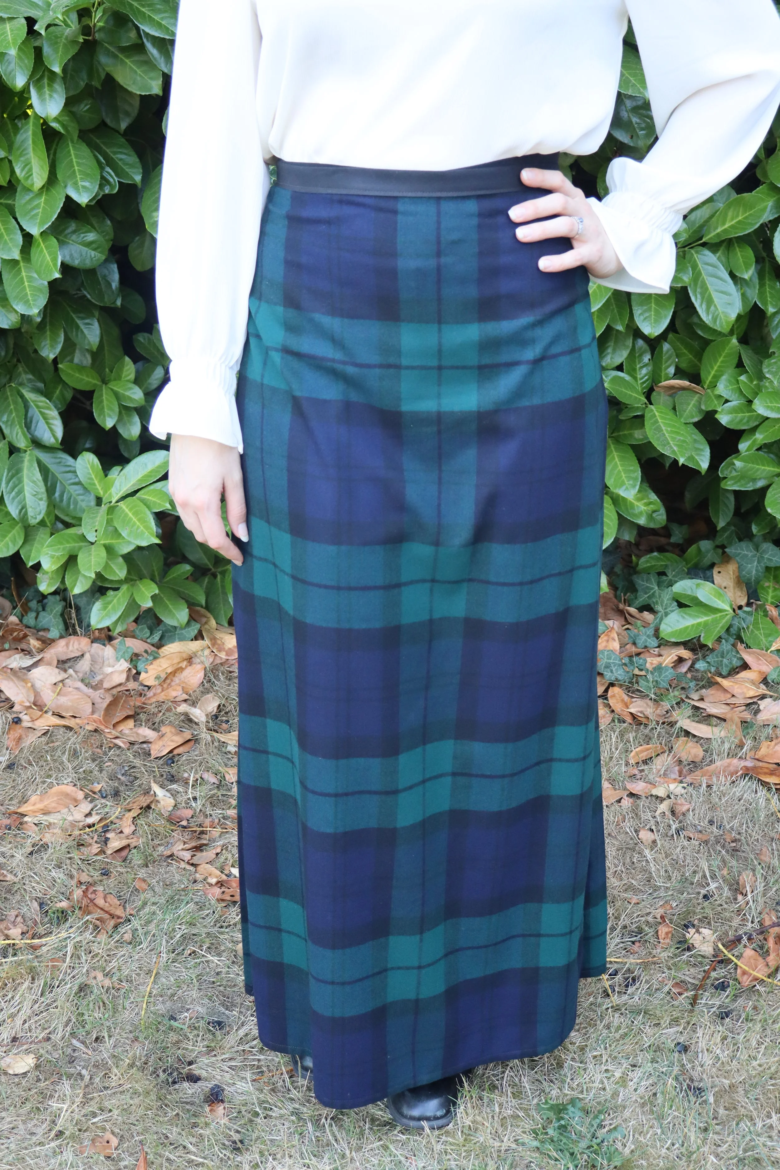 woman wearing a dark green and blue plaid skirt with a white shirt standing in front of green foliage. Garment is medieval inspired, historically influenced clothing.