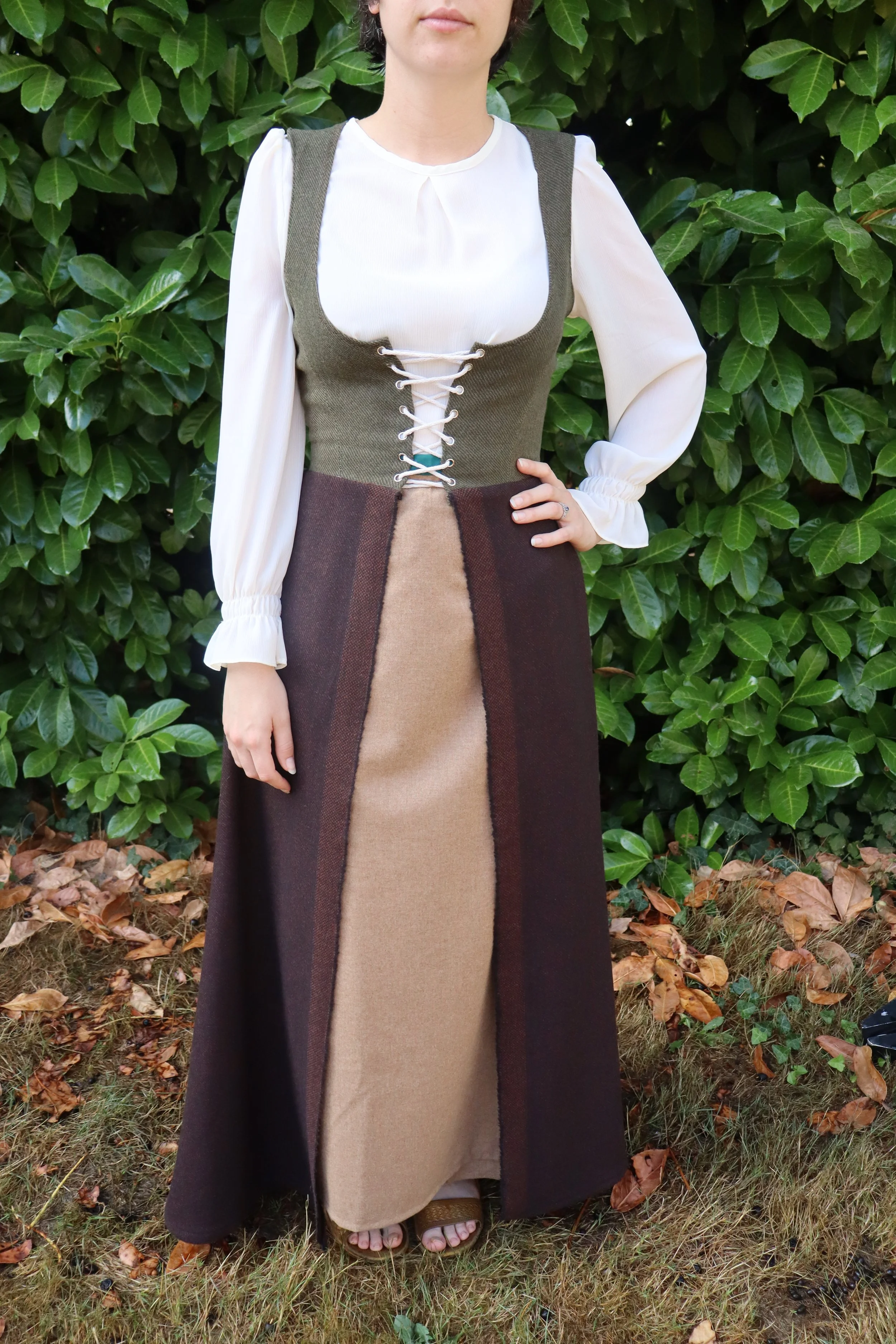 Woman standing outdoors in front of green leafy bushes, wearing a long multicolored dress with a lace-up front detail and a white long-sleeve blouse underneath.