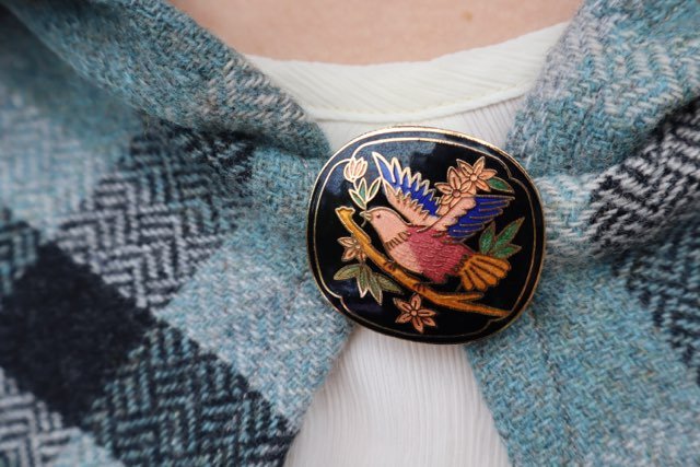Close-up of a cloth with a pin featuring a bird design and floral accents, attached to a 100% wool capelet.