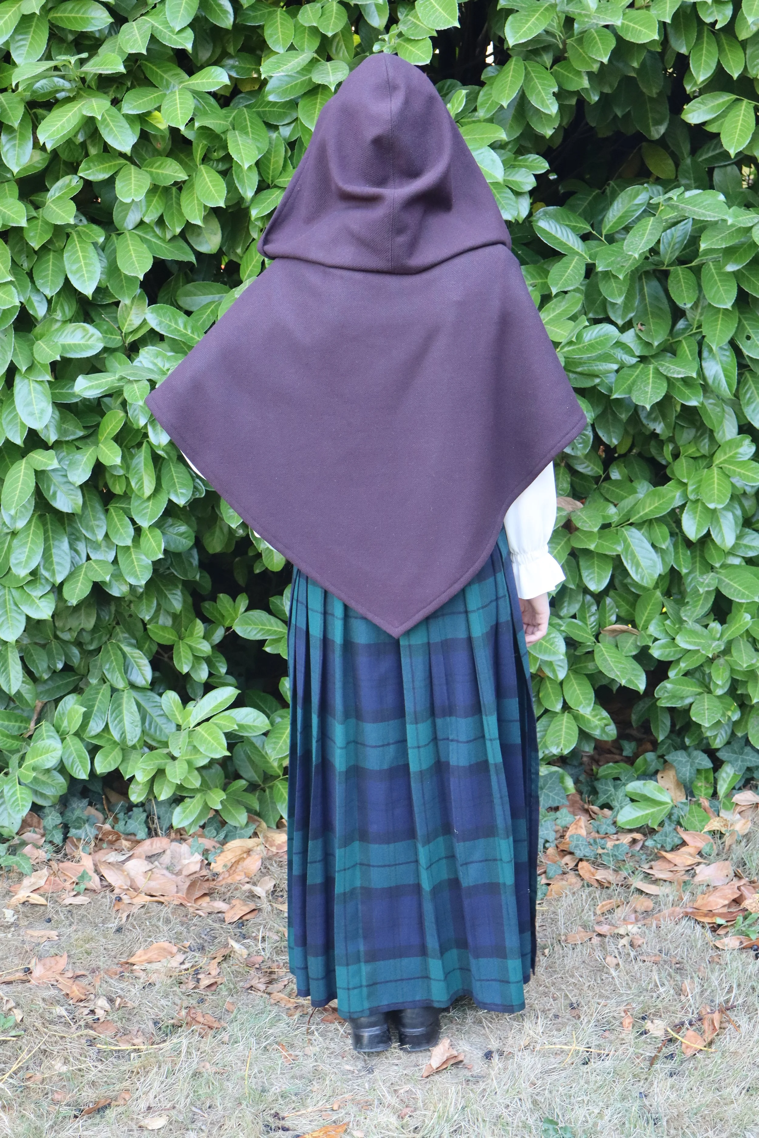 woman in a dark purple capelet/cloak facing away from the camera and standing in front of green foliage.