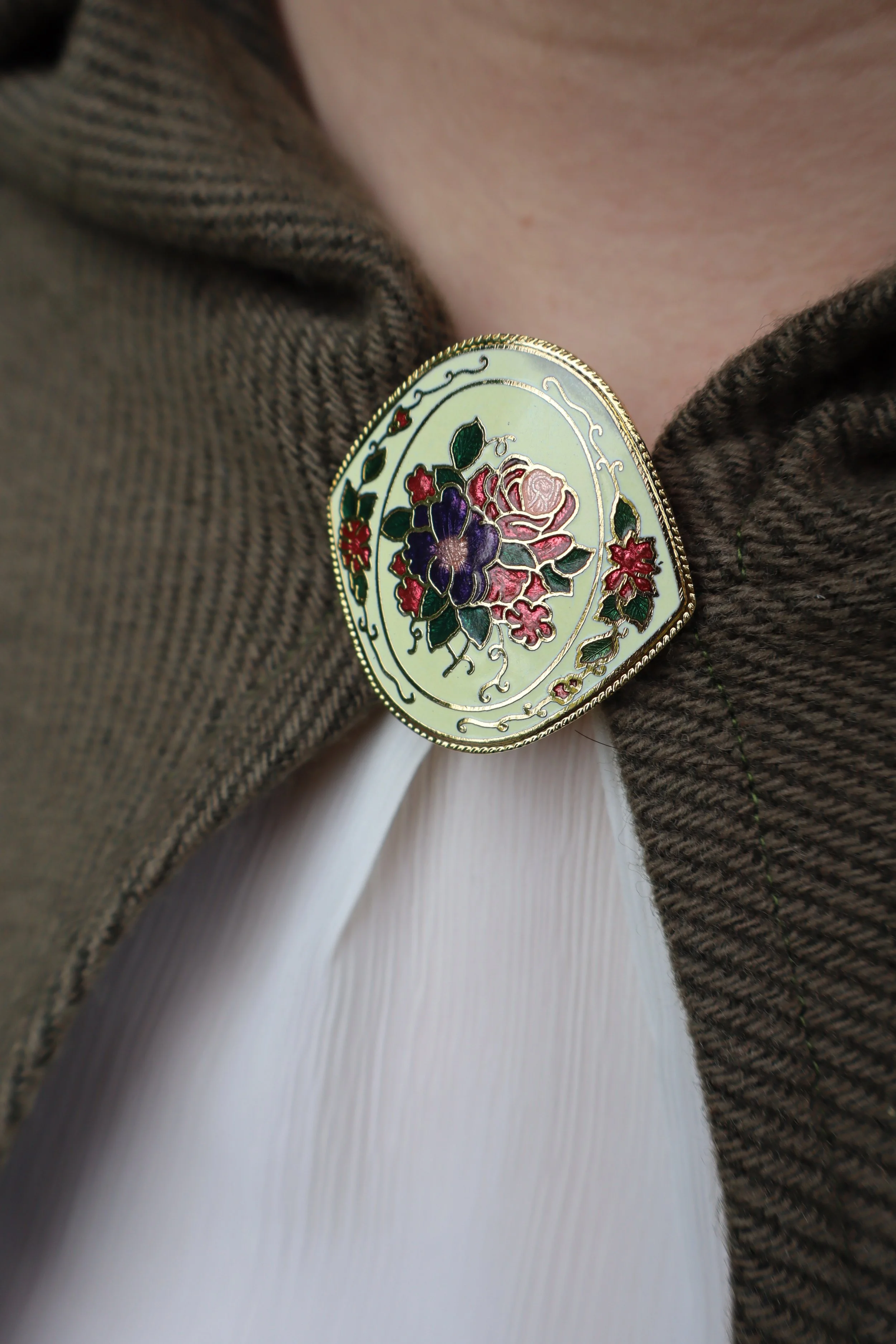 Close up of a floral enamel clasp attached to a green fabric cloak.