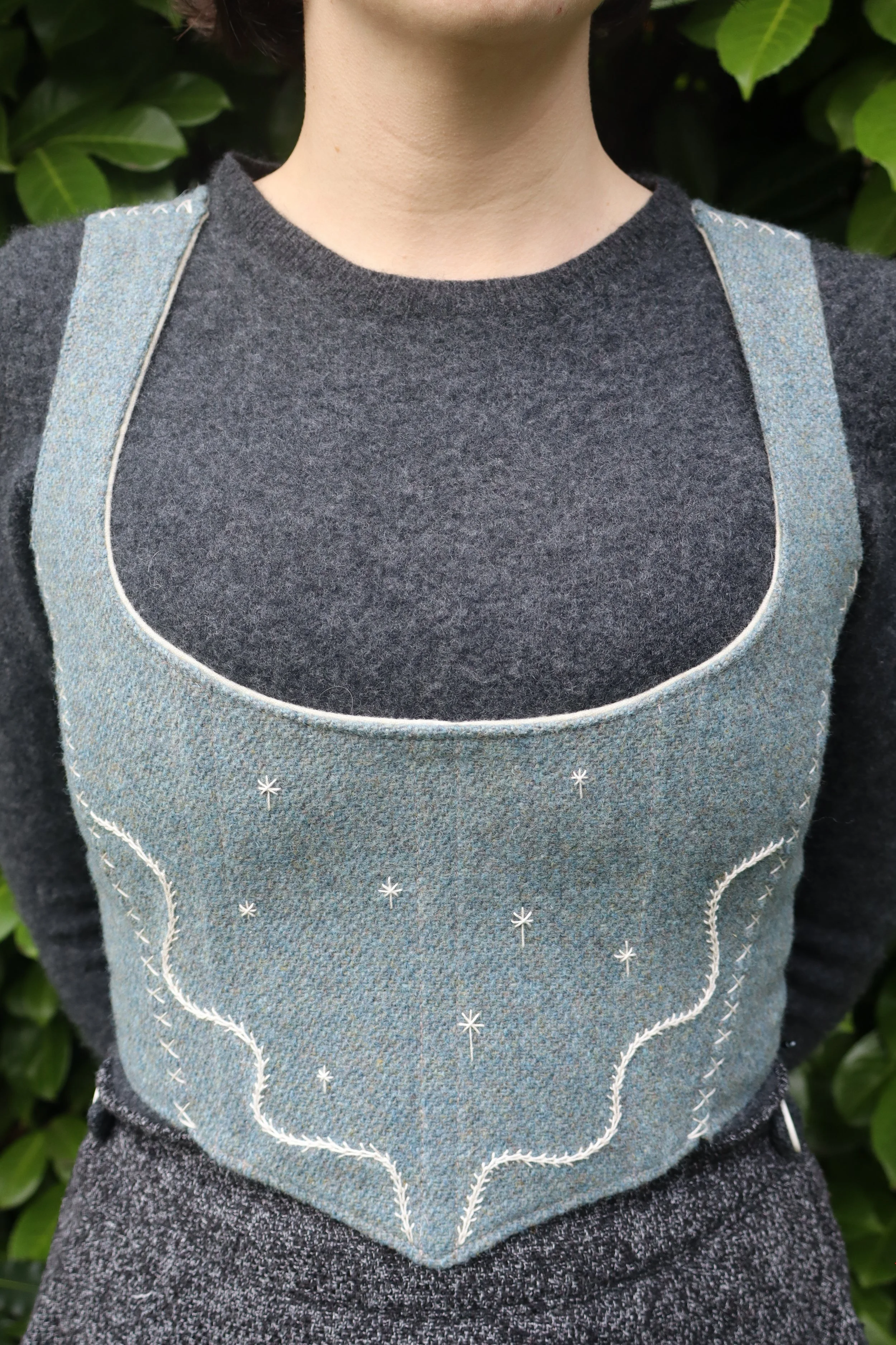 A woman in a grey shirt with a light grey-blue bodice standing in front of vegetation. The bodice has an abstract hand embroidered motif. 