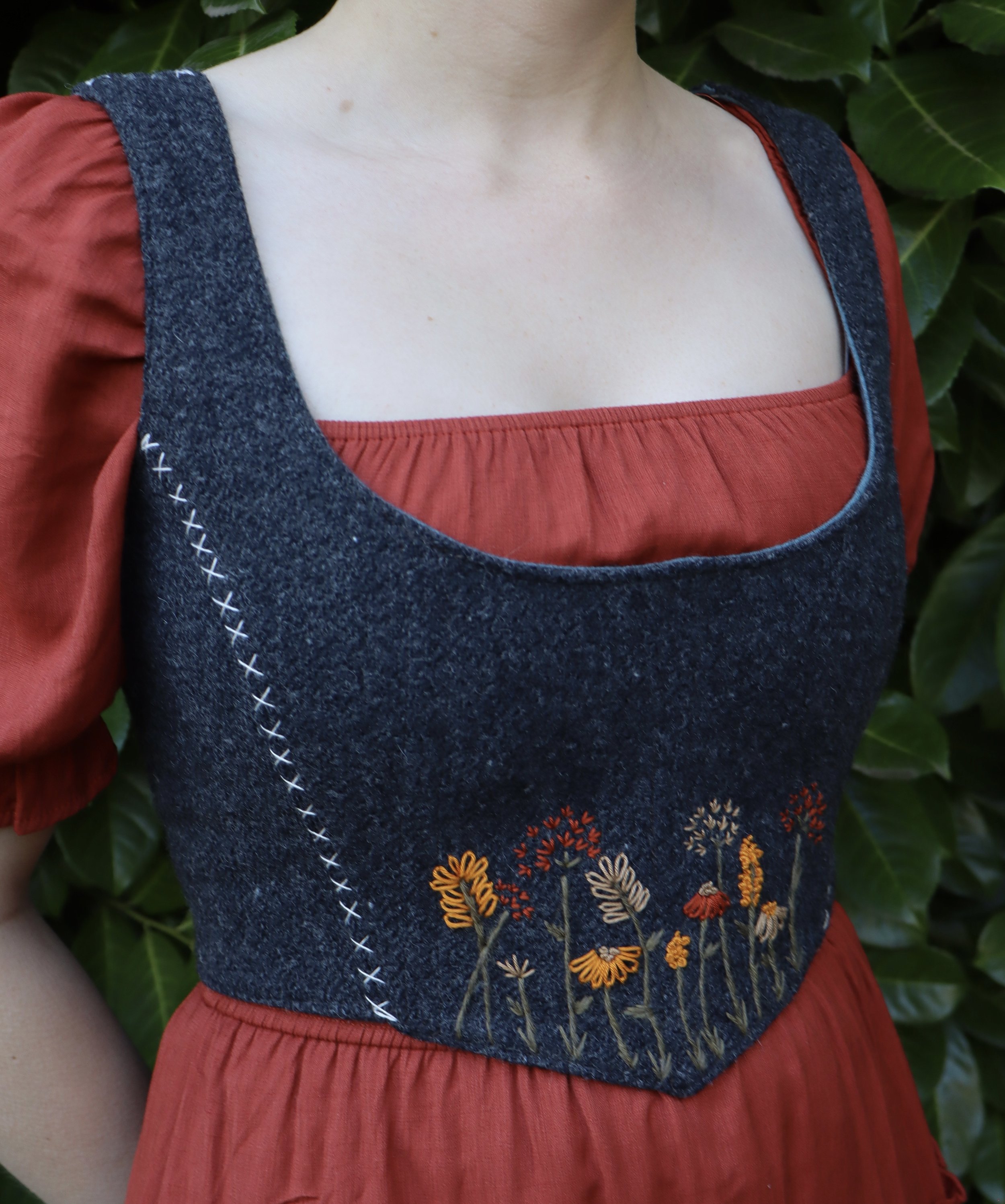 Woman wearing a red undershirt and a grey bodice with an embroidered floral motif.
