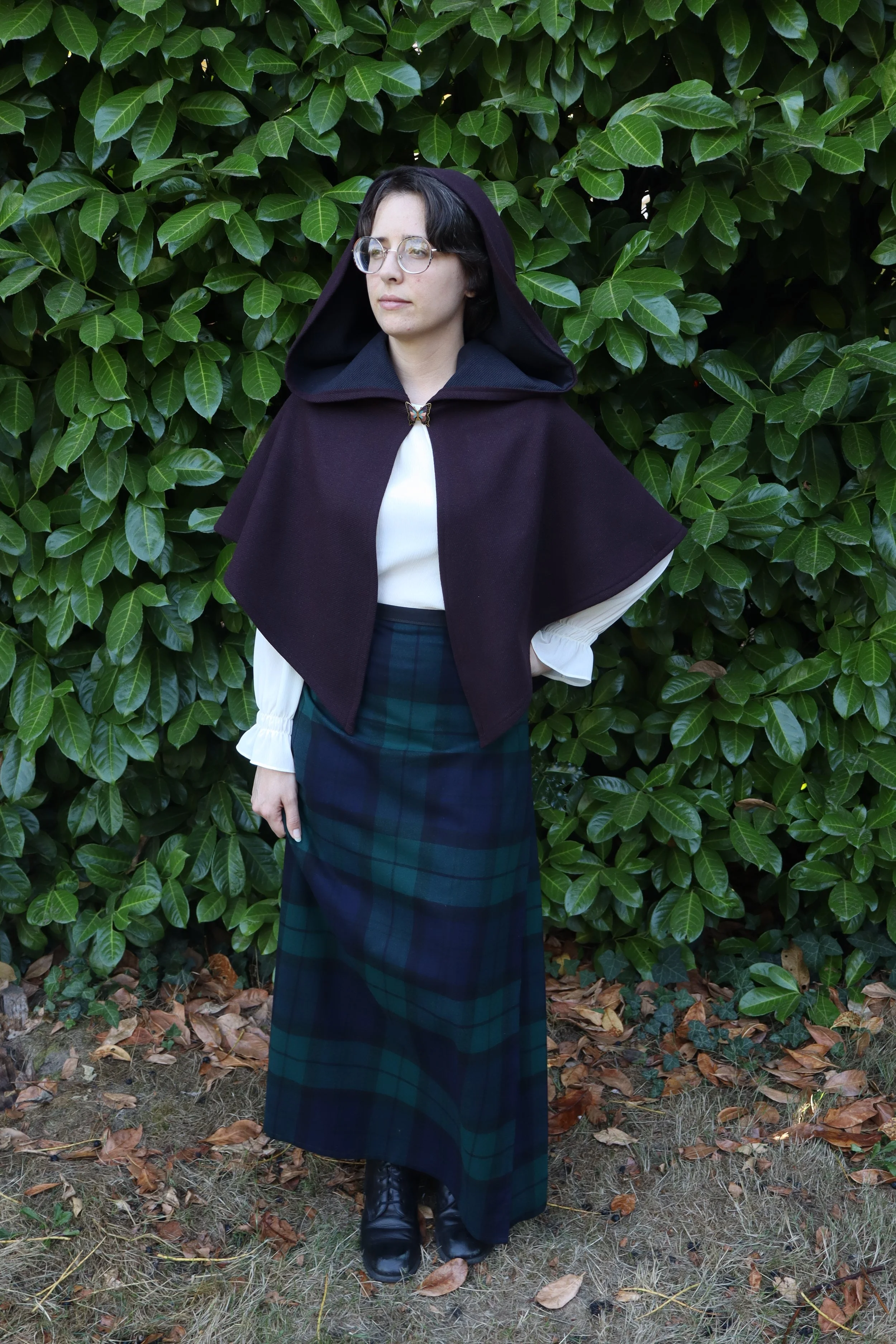 woman with glasses in a dark purple capelet/cloak  and a white shirt standing in front of green foliage.