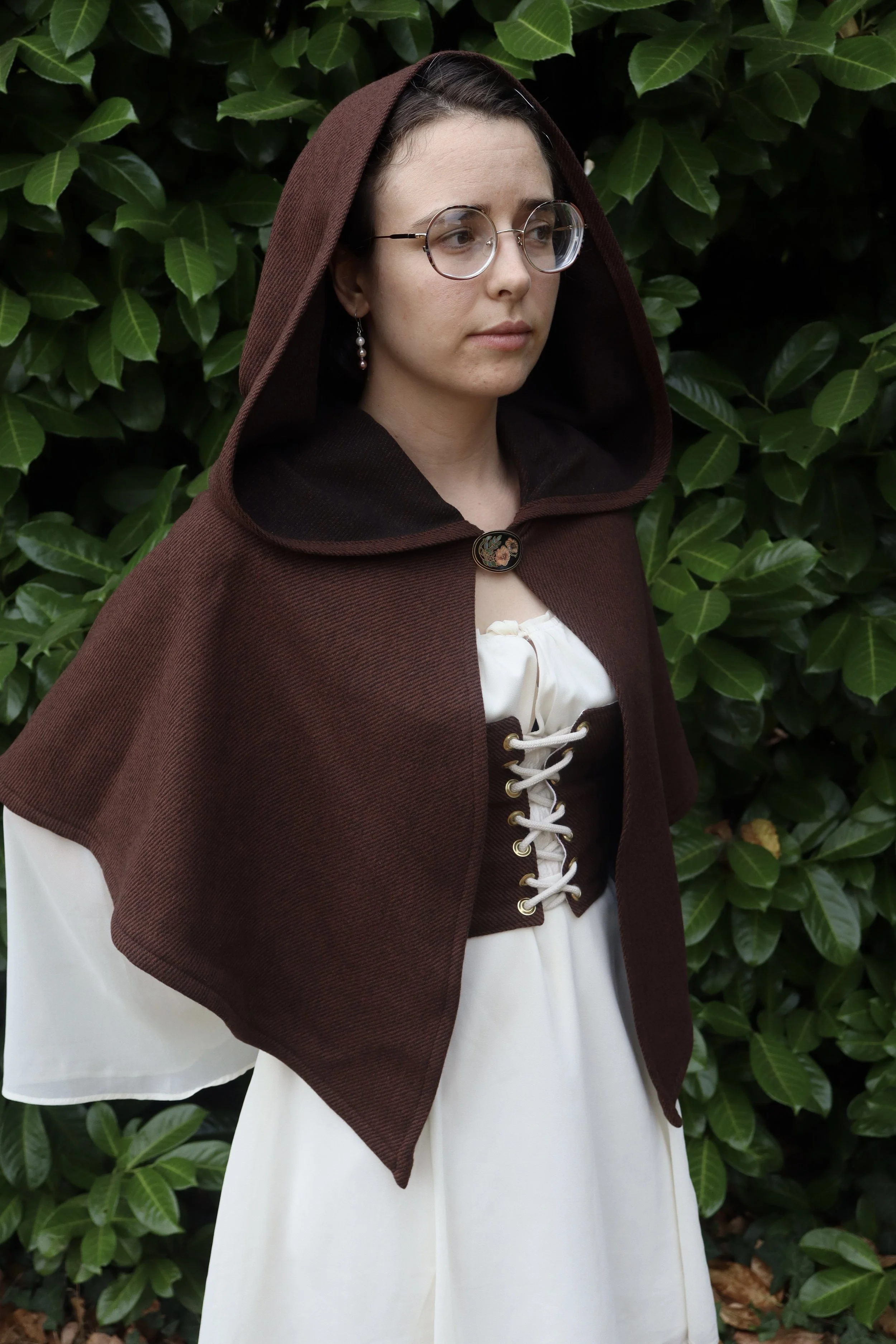 woman with glasses in a brown capelet/cloak  and a white shirt standing in front of green foliage.