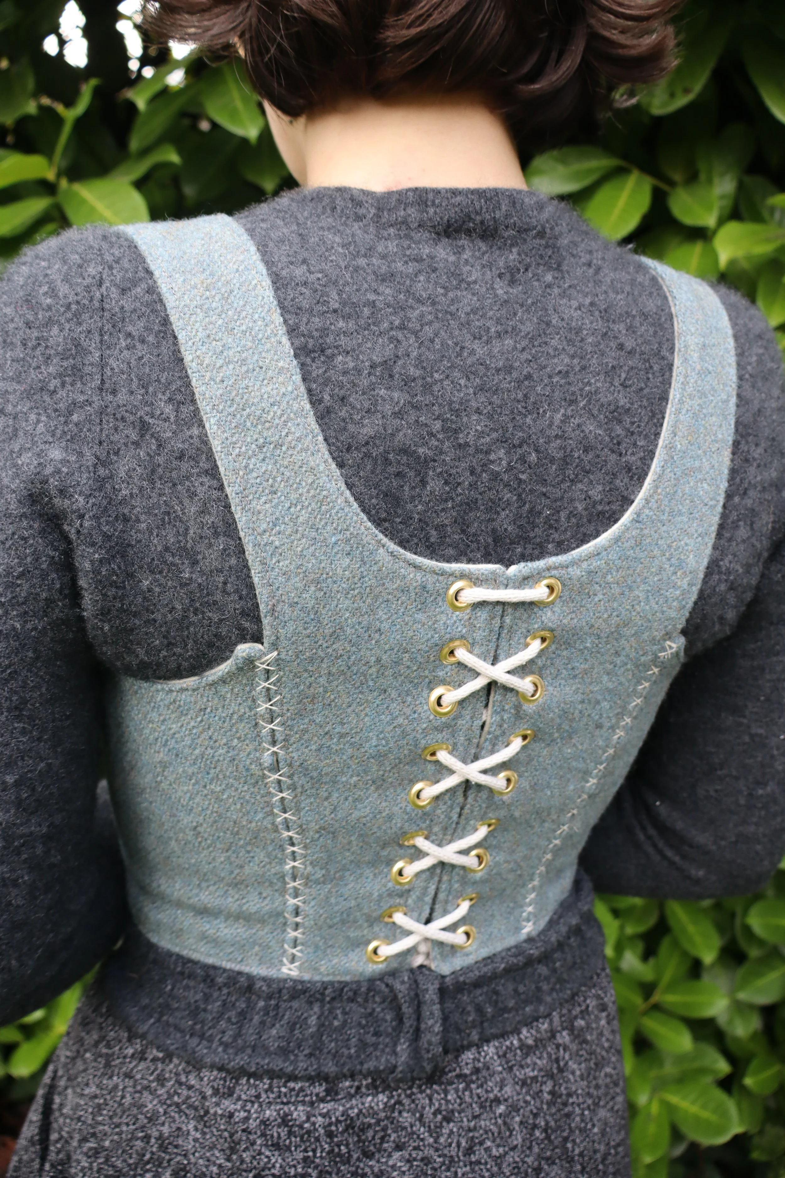 Woman with back facing camera wearing a grey shirt with a light grey-blue bodice and lacing. Standing in front of a green bush