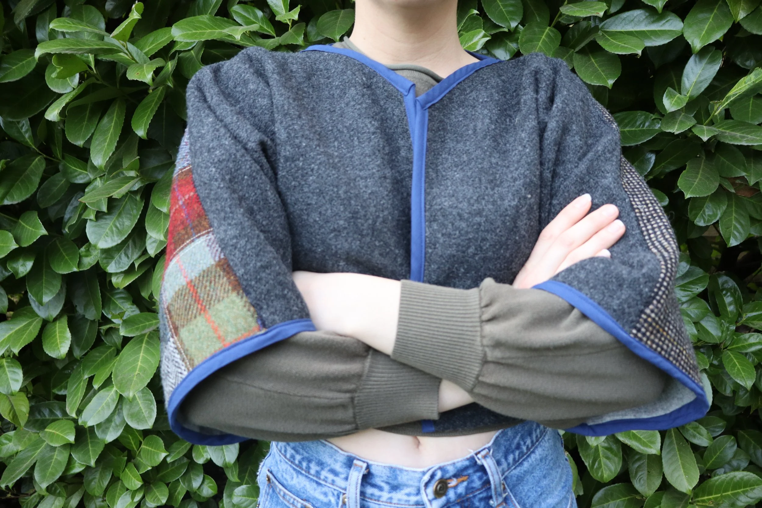 Person wearing a dark gray hoodie with plaid and mesh patches on sleeves, blue trim, and an olive green long-sleeve shirt underneath standing in front of green leafy bushes with arms crossed.