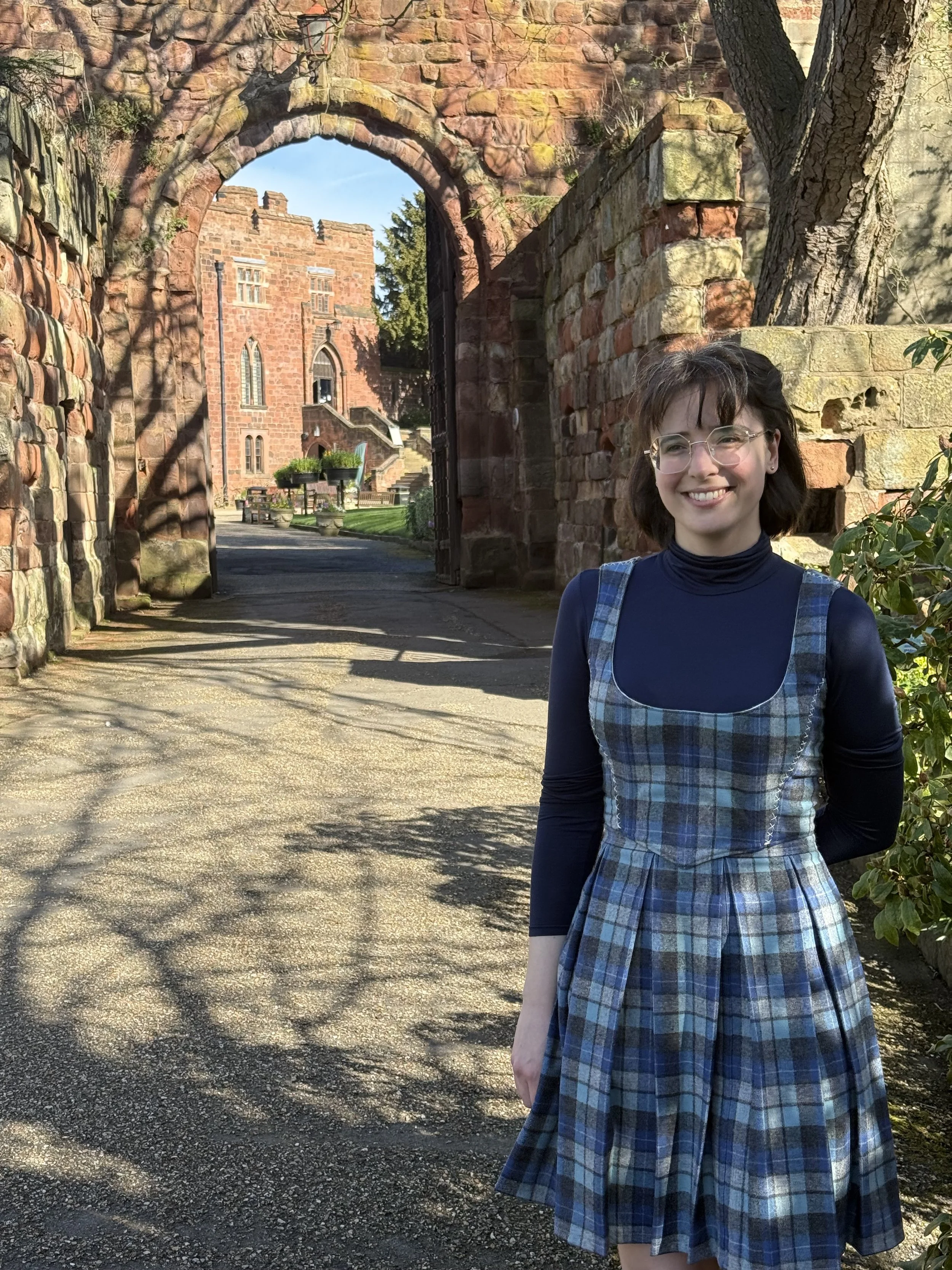 woman in blue plaid dresss standing in front of large red stone castle gate
