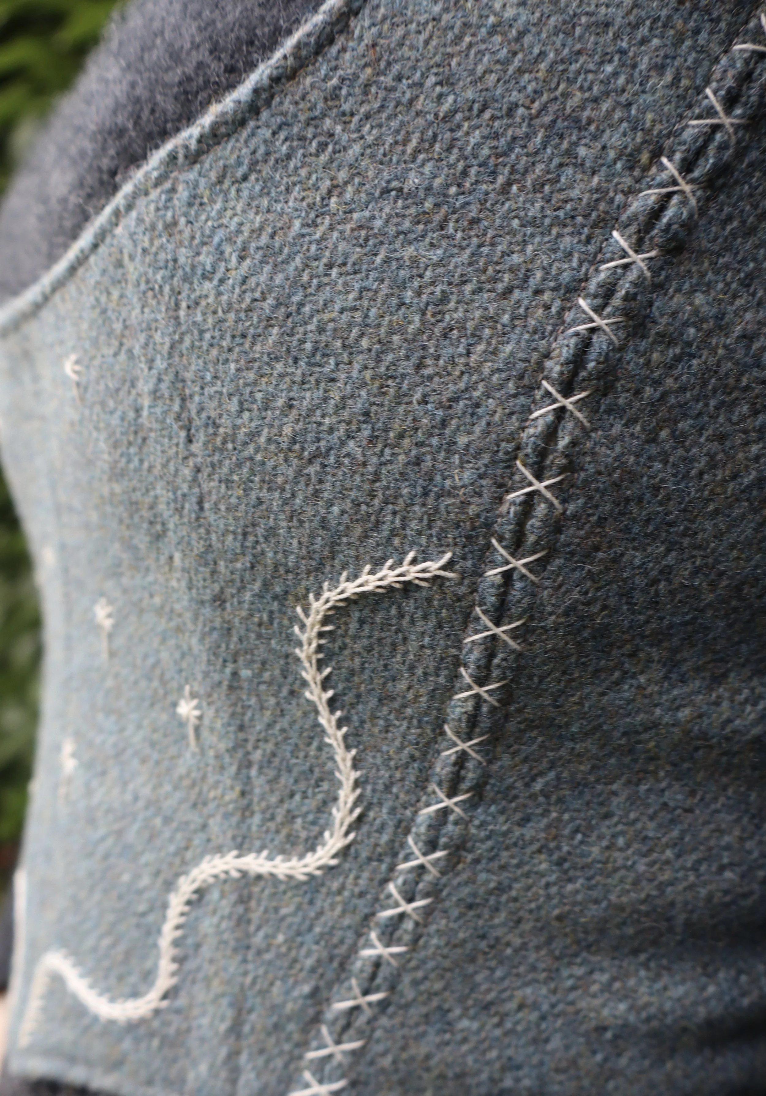 upclose of a Woman wearing a grey shirt with a light grey-blue bodice with a abstract embroidered motif. Standing in front of a green bush