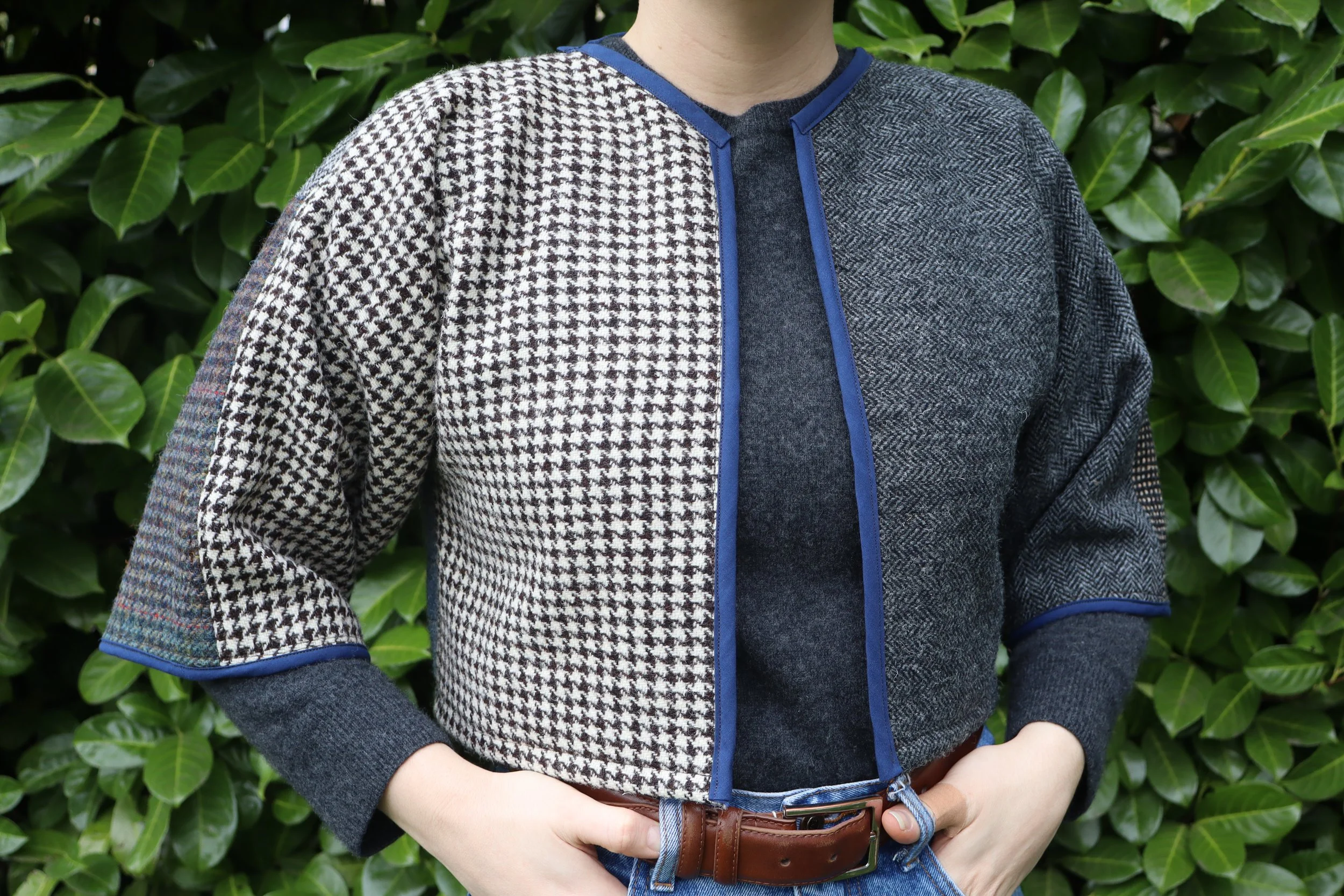 Person wearing a patchwork-style jacket with different patterns, standing in front of green leafy bushes.