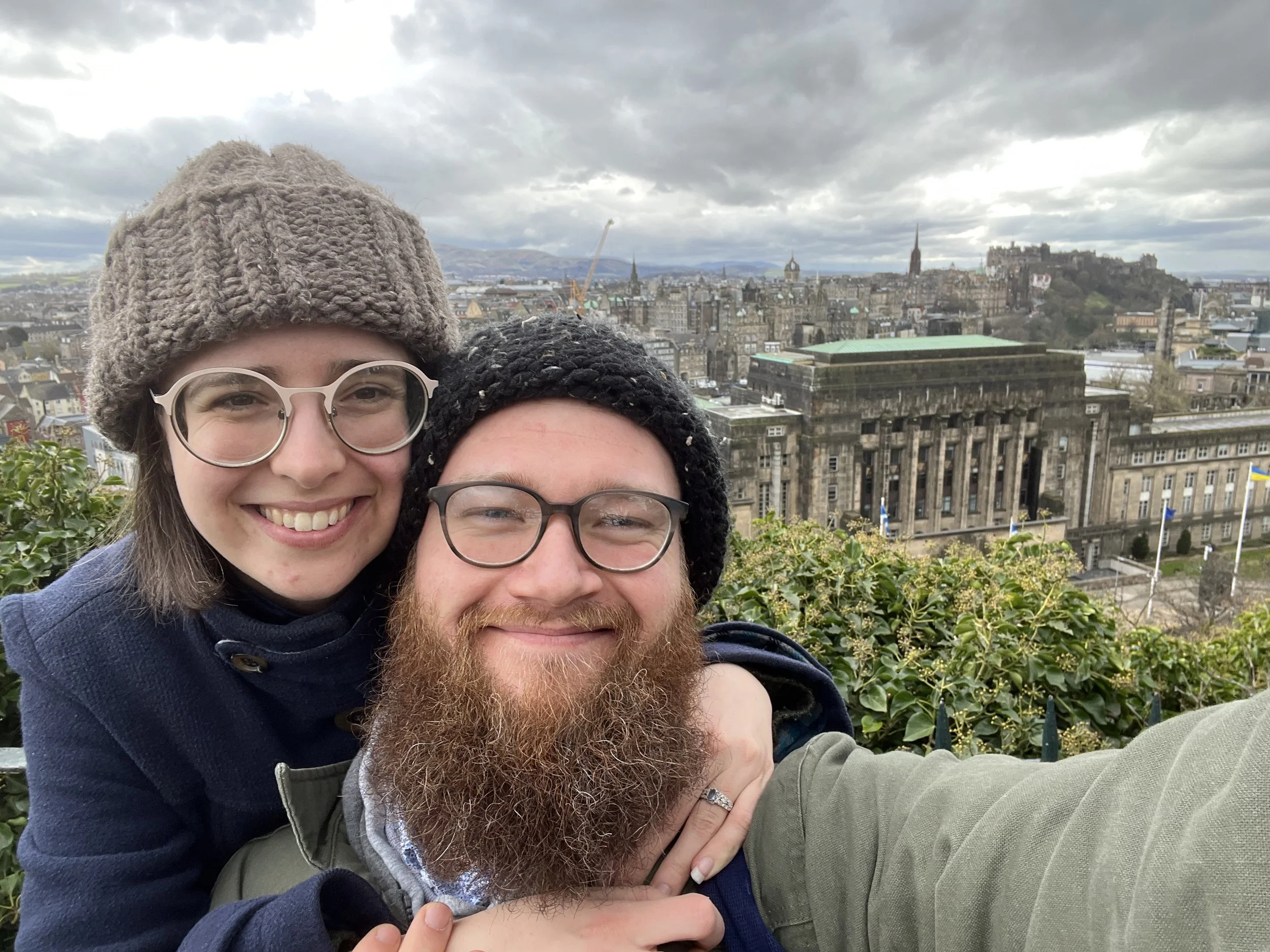 A smiling couple taking a selfie with a cityscape in the background, including historic buildings and cloudy sky.