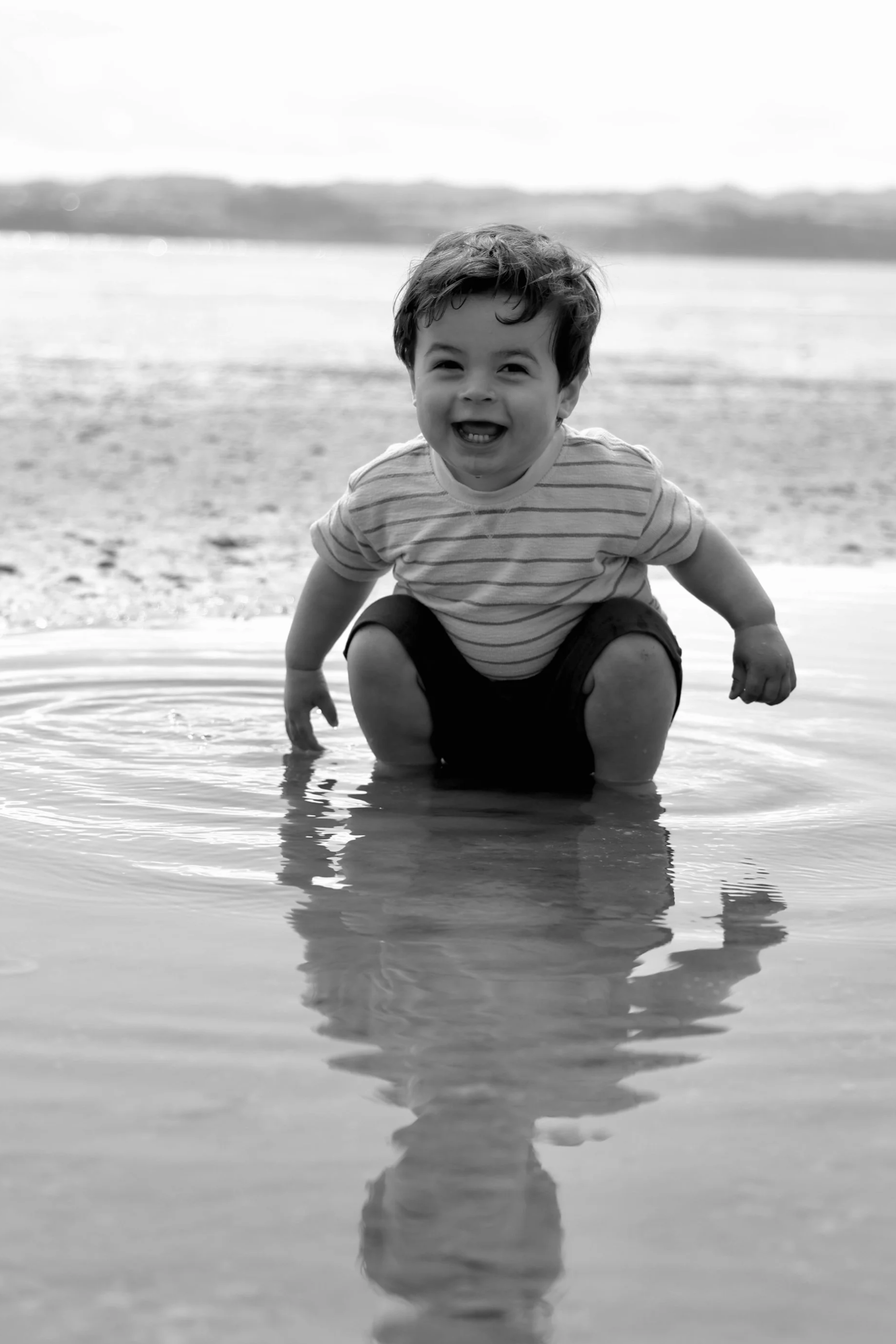 A happy young boy playing in the shallow water at the beach, wearing a striped t-shirt and shorts, with a joyful expression and reflection on the water.