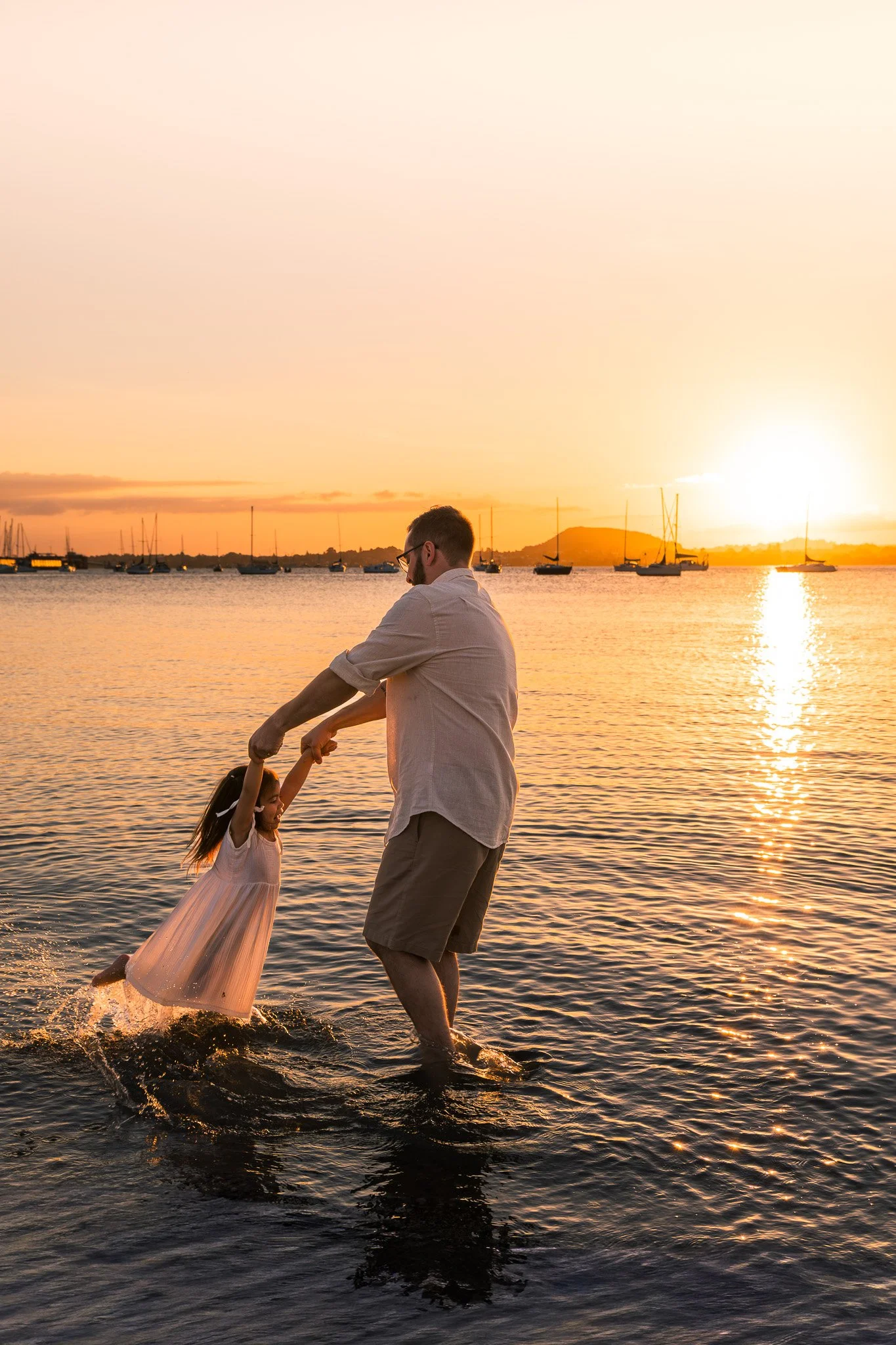 A man and young girl playing in water during sunset, with boats on the water and a distant mountain in the background.