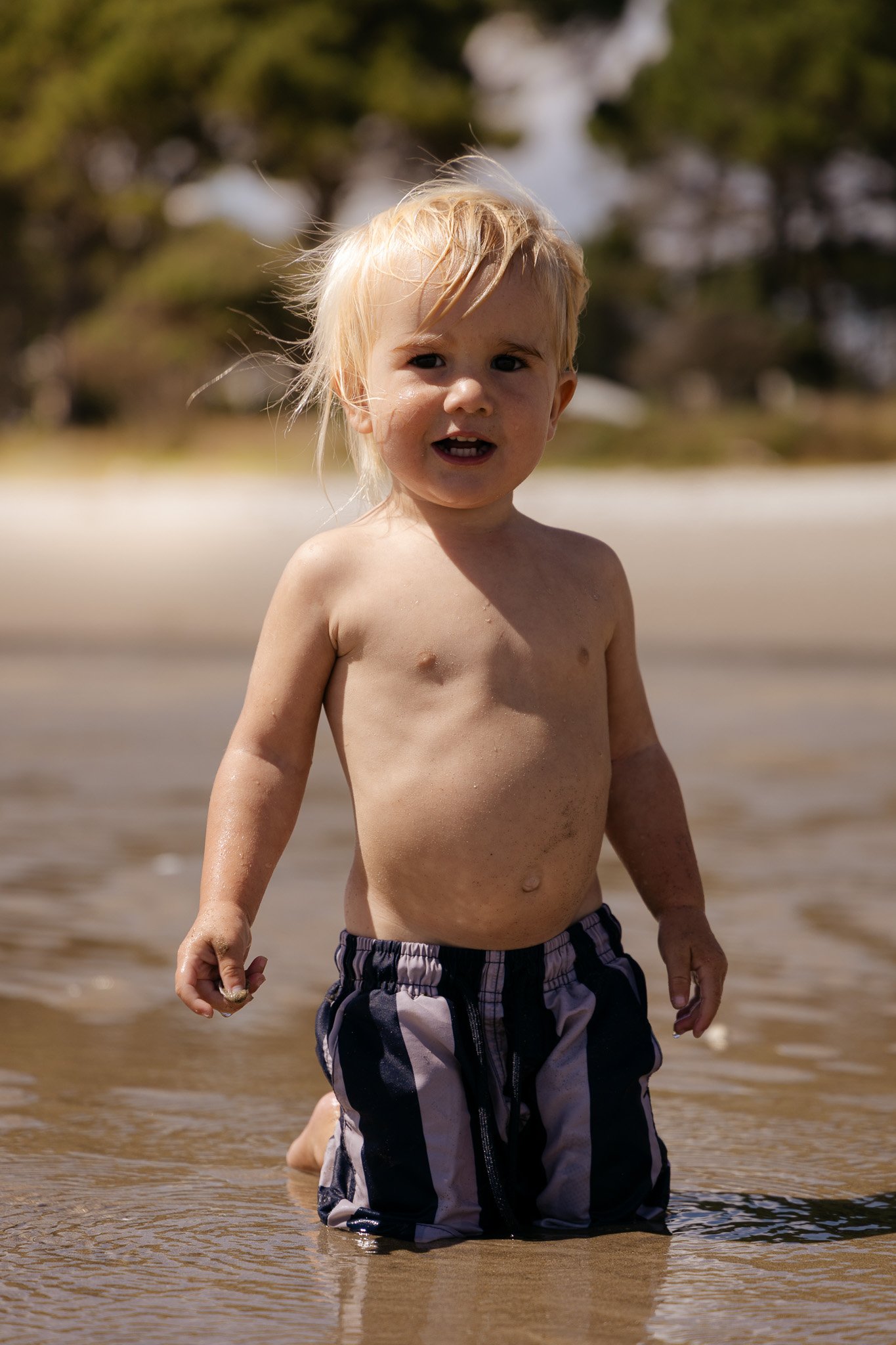 A young blond boy standing in shallow water at the beach, smiling and looking at the camera.