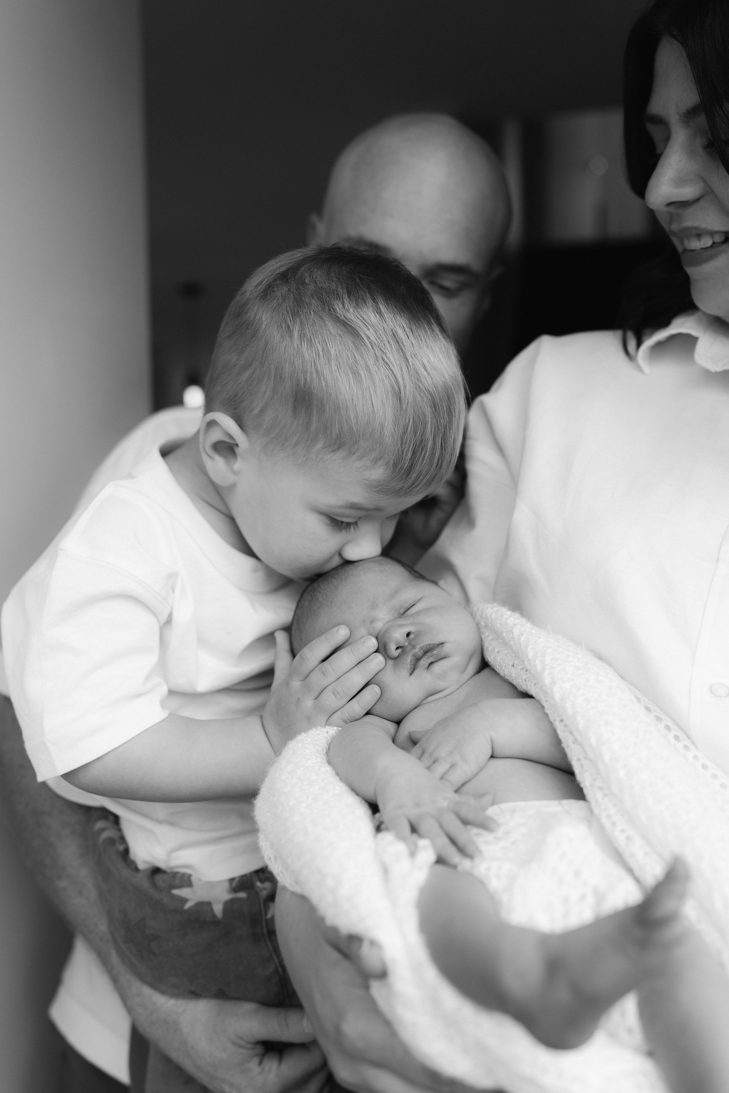 older brother kissing newborns forehead with family in his house