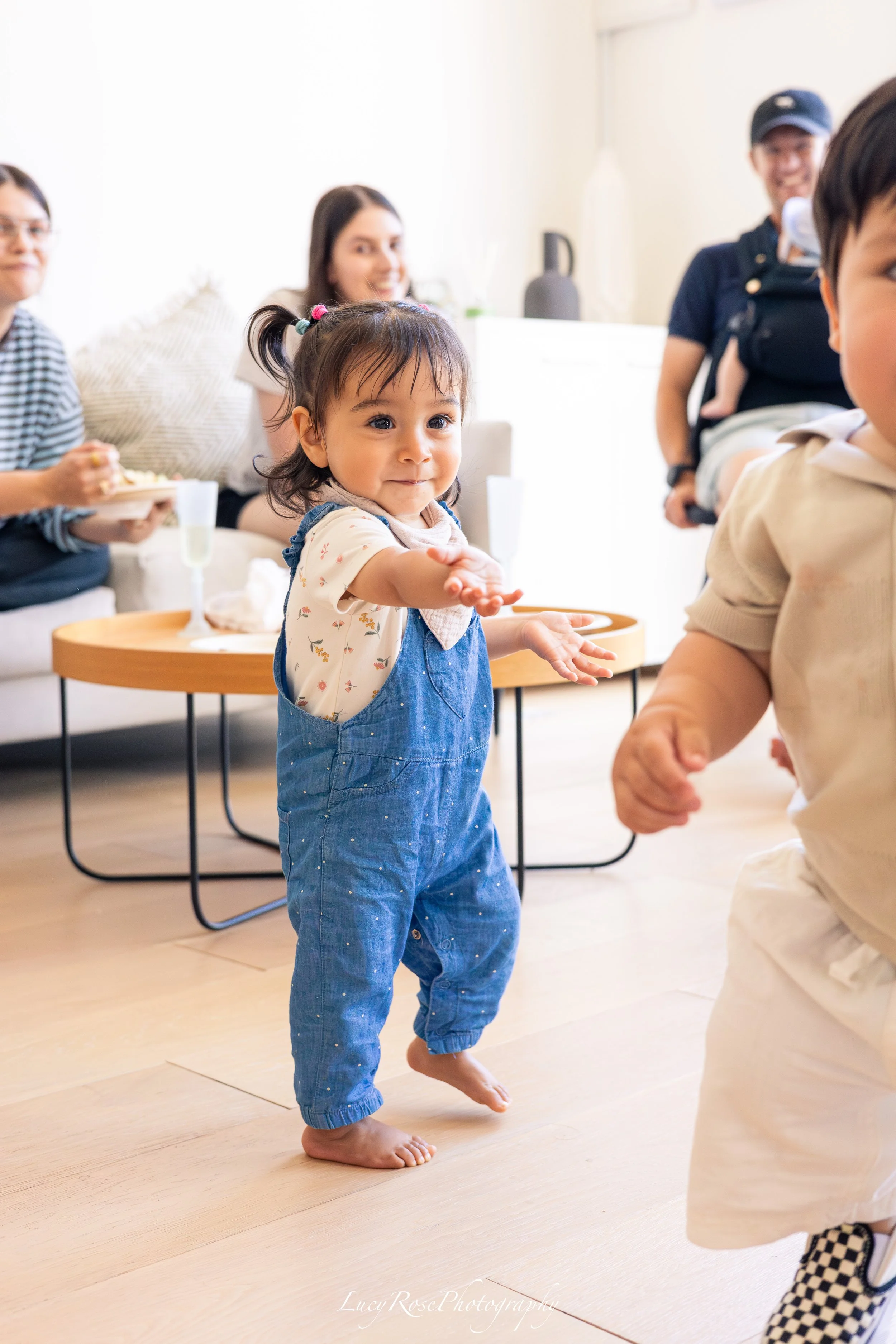 Young girl with dark hair in pigtails, wearing denim overalls and a floral shirt, standing barefoot on a wooden floor, clapping and smiling amid other children and adults in a bright indoor setting.