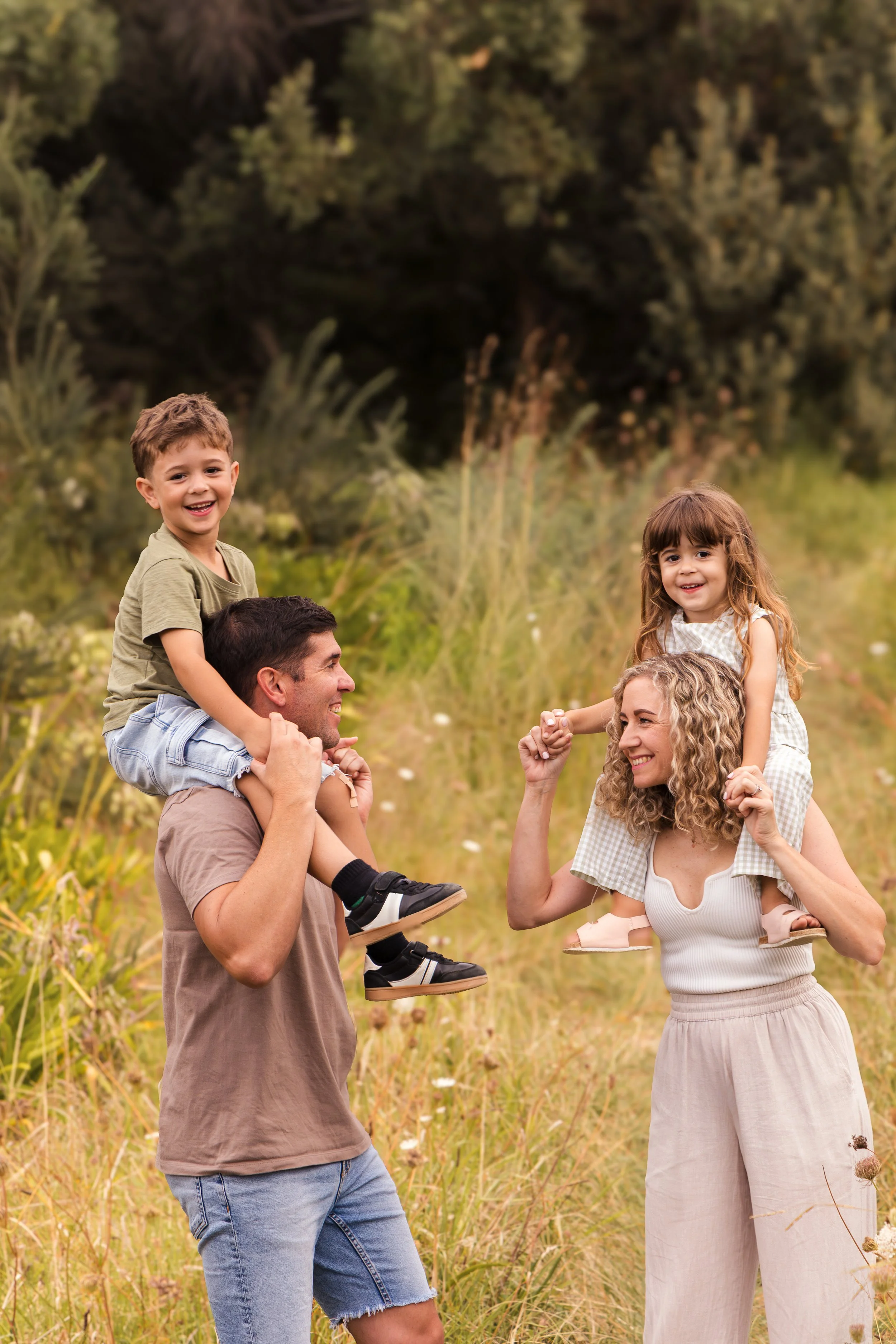 Family of four, two children and two adults, spending fun time outdoors in nature, with the kids sitting on the parents' shoulders and smiling.
