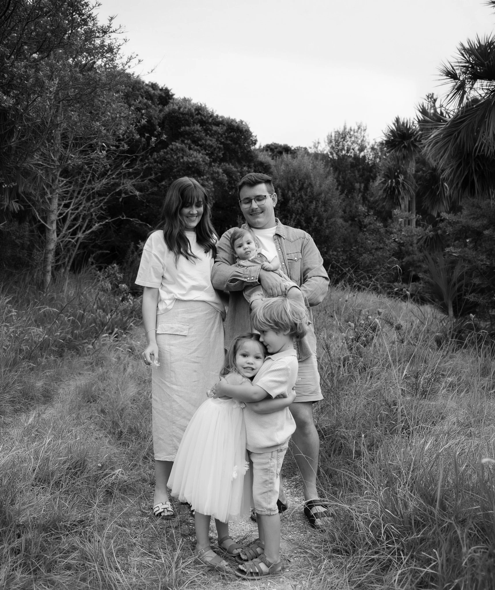 A black-and-white photograph of a family of five standing on a grassy trail surrounded by trees and bushes, smiling and embracing each other.
