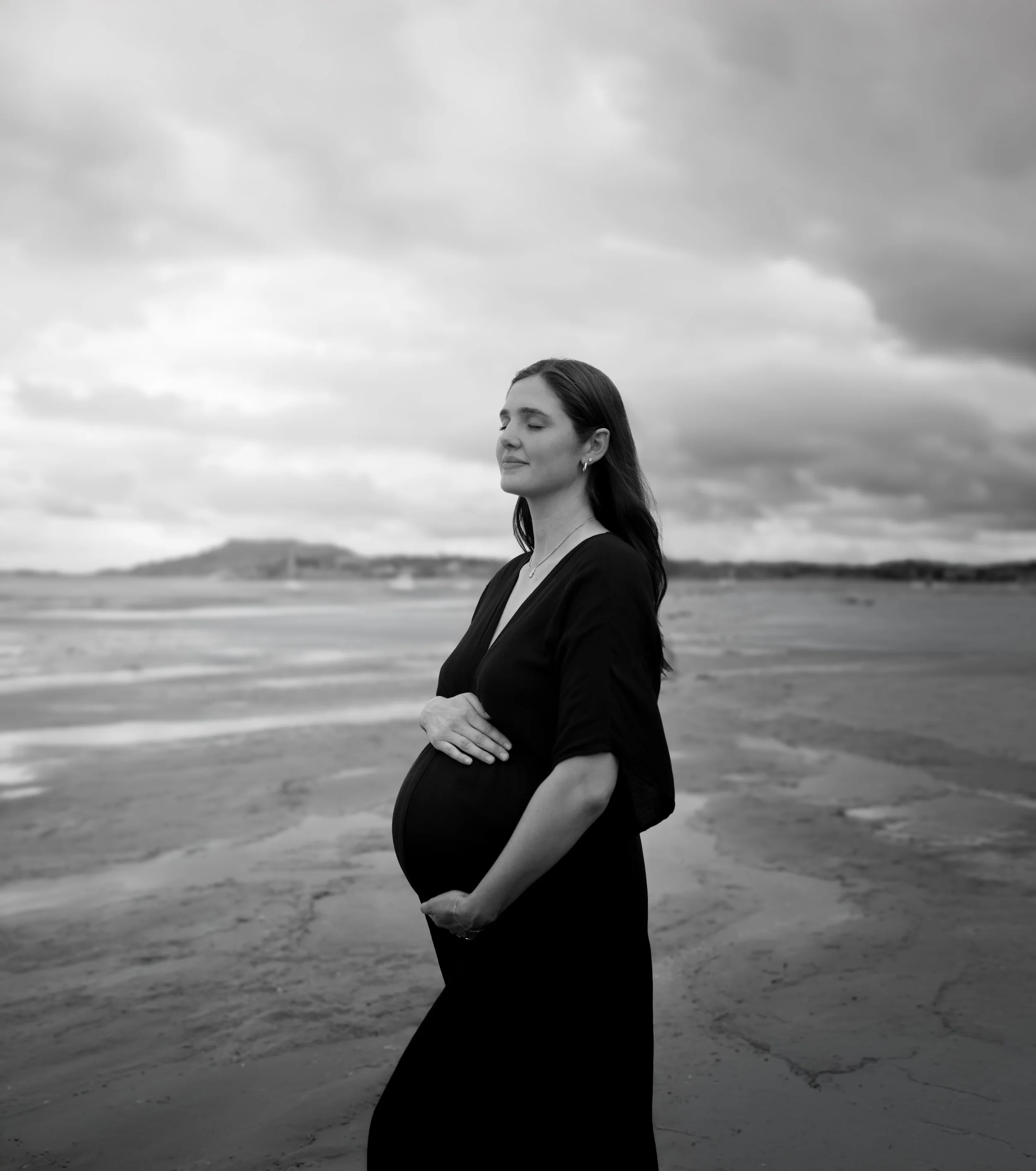 Black and white photo of a pregnant woman standing on a beach with her eyes closed, gently holding her belly with one hand and the other hand resting on top of it, under a cloudy sky.