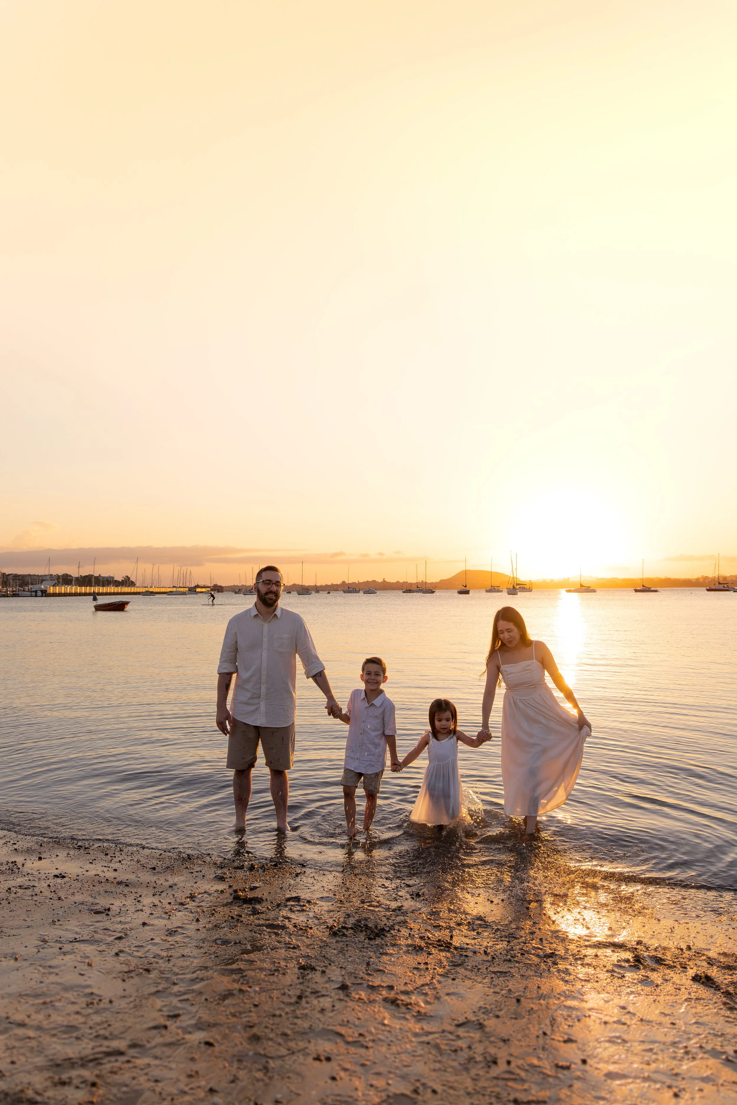 A family of four holding hands and standing in the shallow water at the beach during sunset.