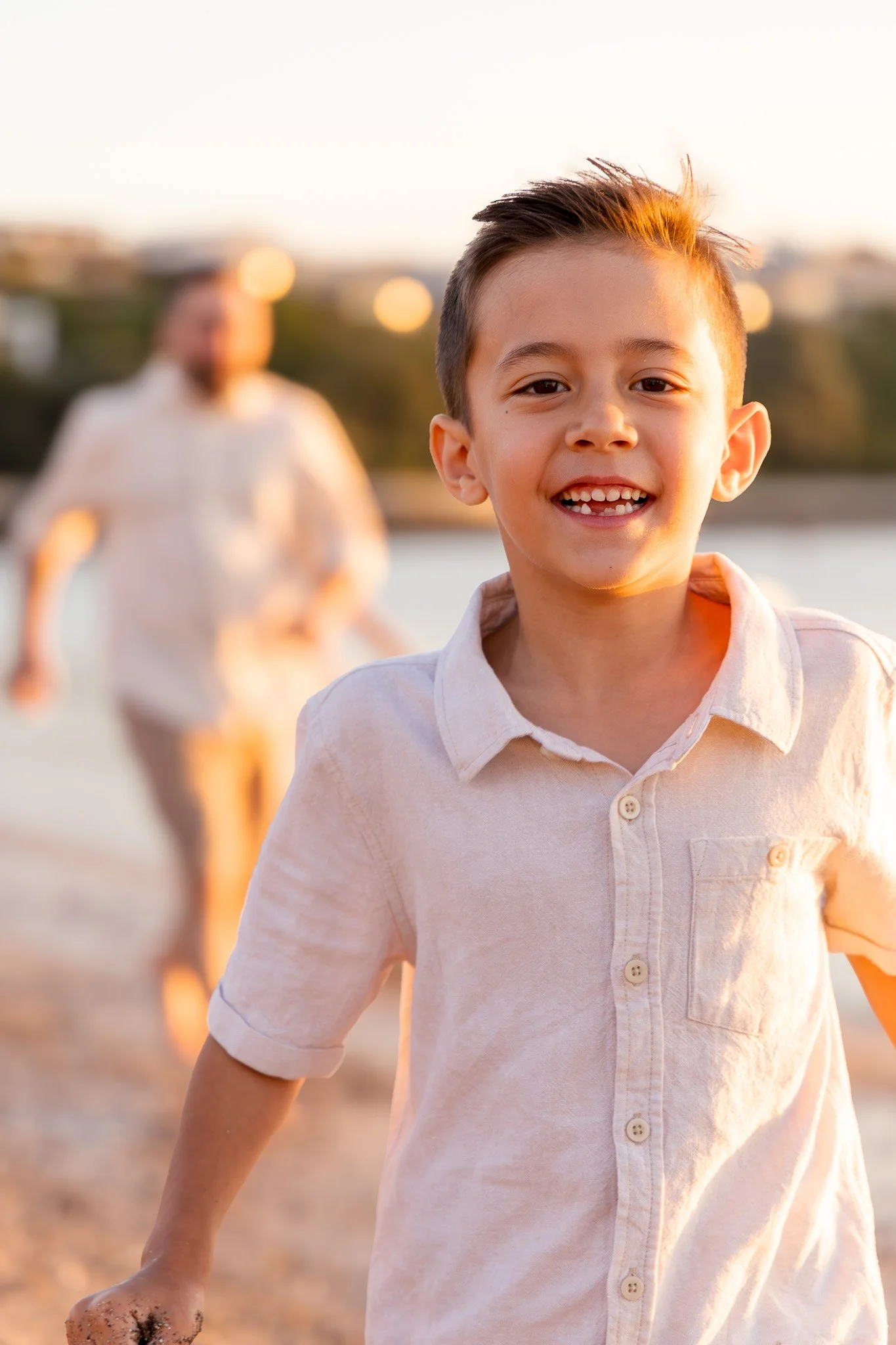 A young boy smiling and running outdoors at sunset with a woman blurred in the background.