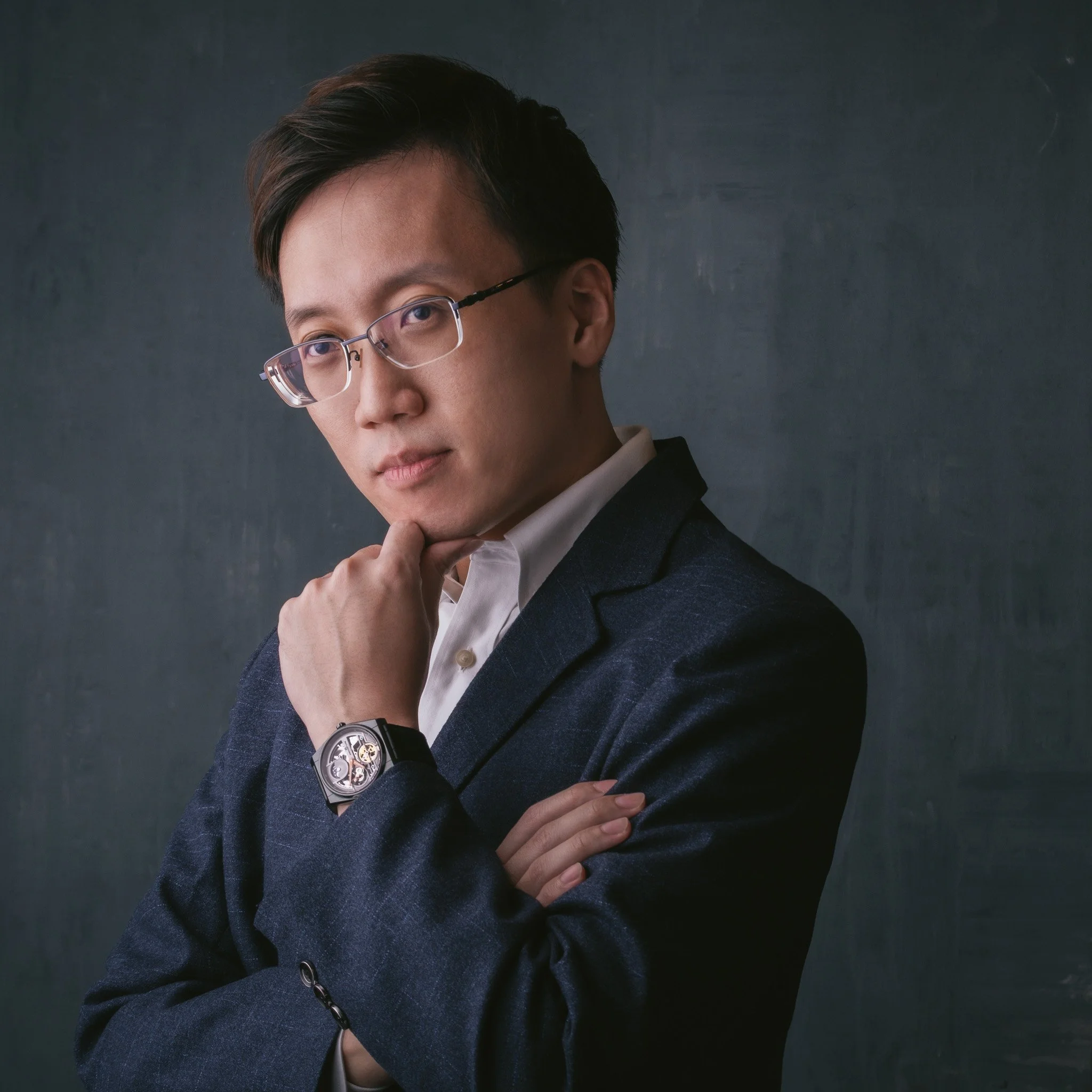 A young man with short dark hair and glasses wearing a dark blue blazer and white shirt, posing against a dark background.