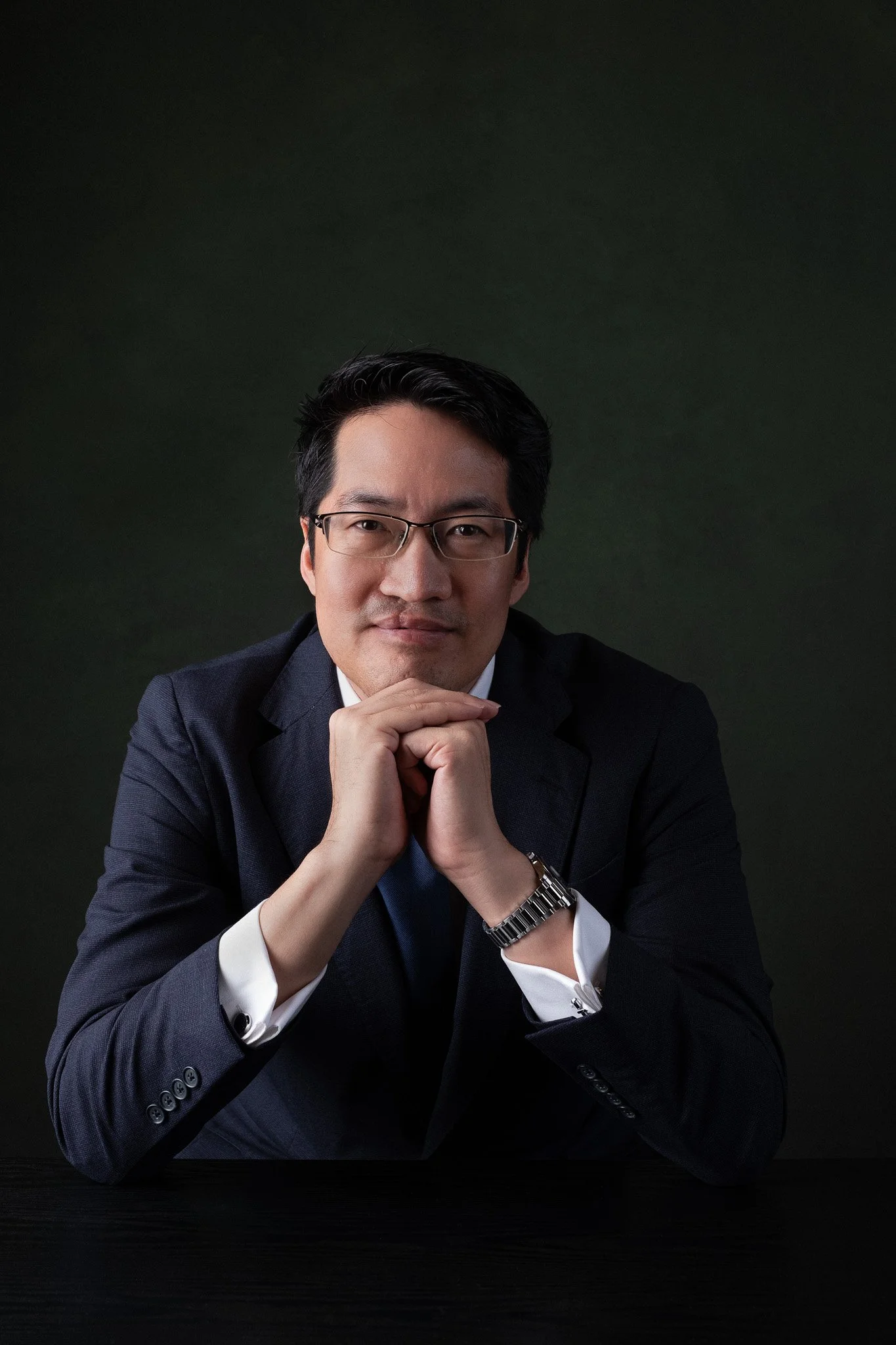 Executive portrait for Lawrence Chong, CEO of Consulus Group, Singapore. He is seated, facing the camera with hands below his chin. Shot against a dark background.