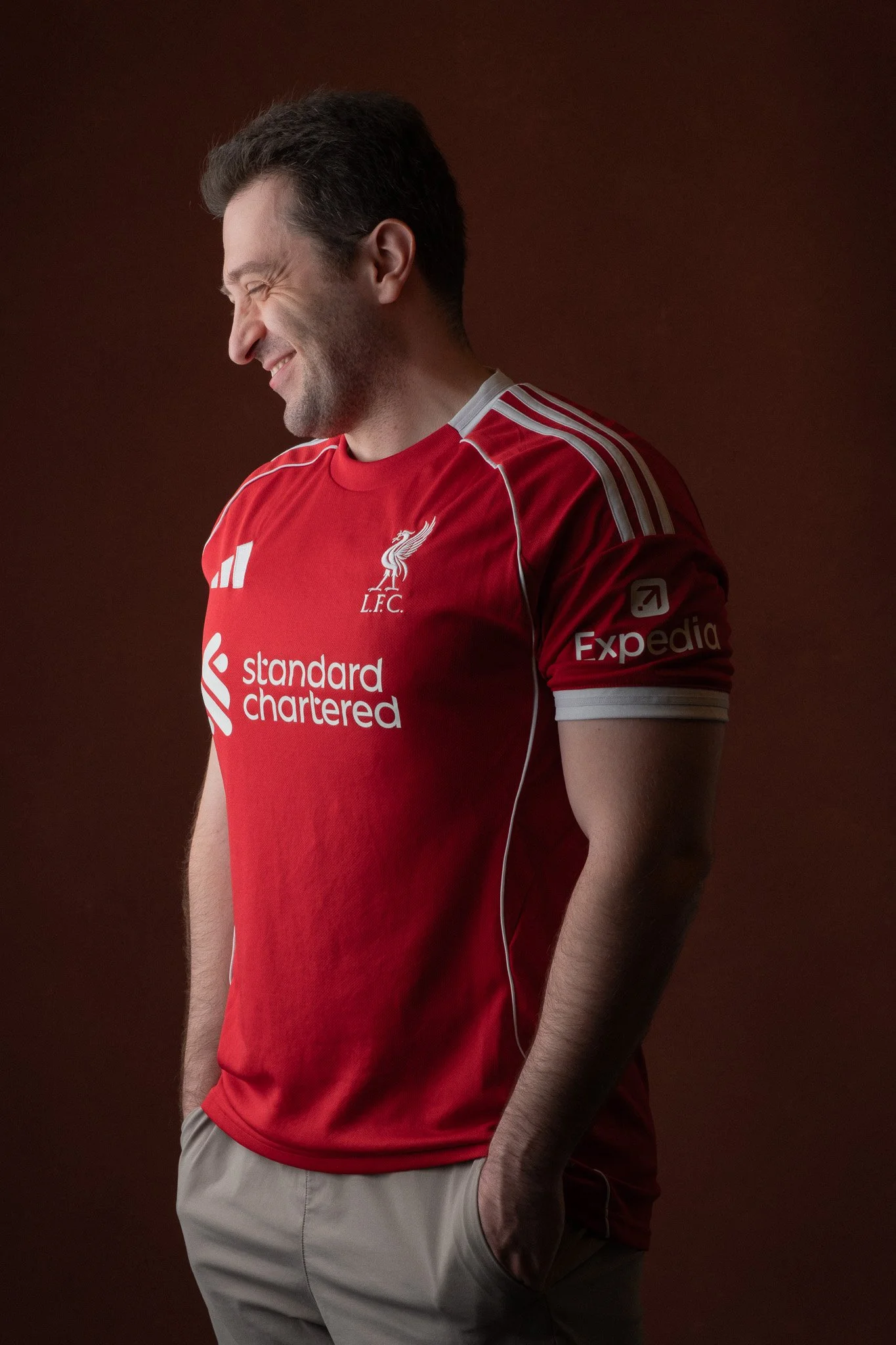 Side profile of a man smiling wearing a red Liverpool football jersey against a dark background.