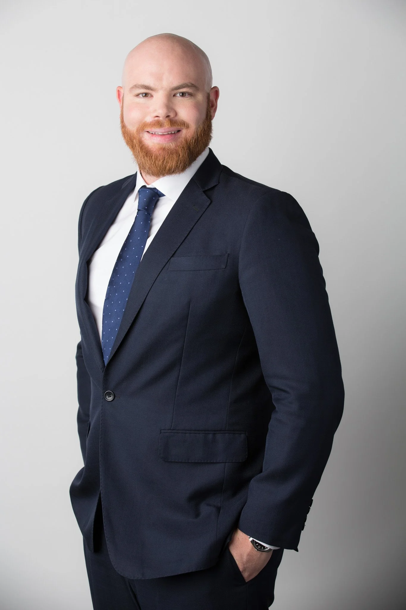 A man with a bald head and red beard in a dark suit, white shirt, and blue tie with small white dots, standing against a plain white background. He has his left hand in his pocket and is smiling.