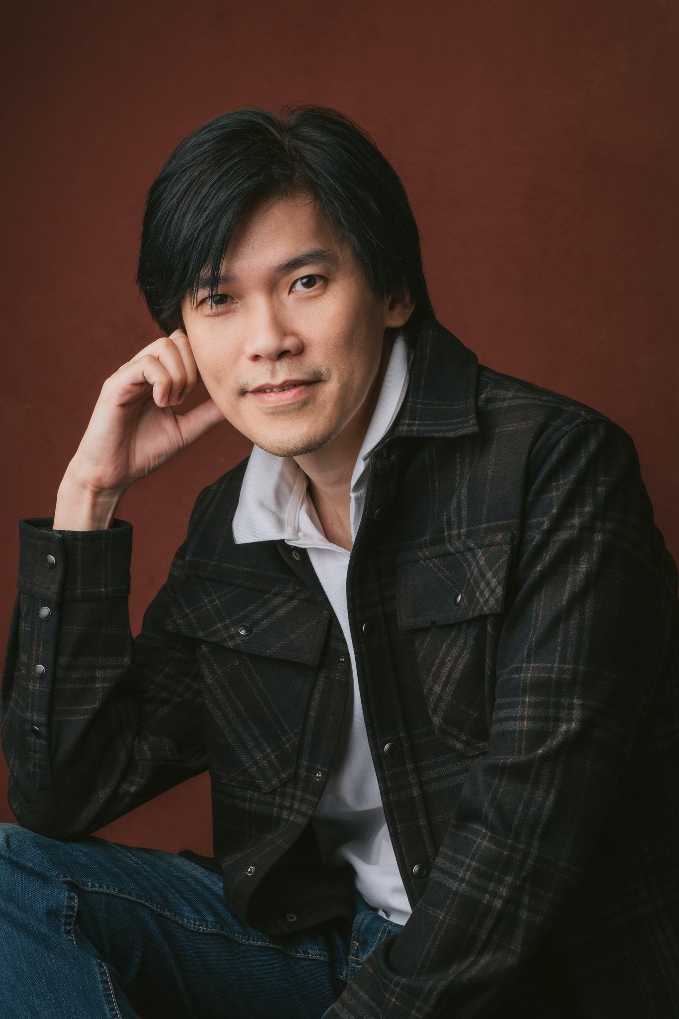 A man with black hair poses for a portrait against a brown background. He is wearing a black plaid jacket over a white shirt and blue jeans, with his right hand resting near his face.