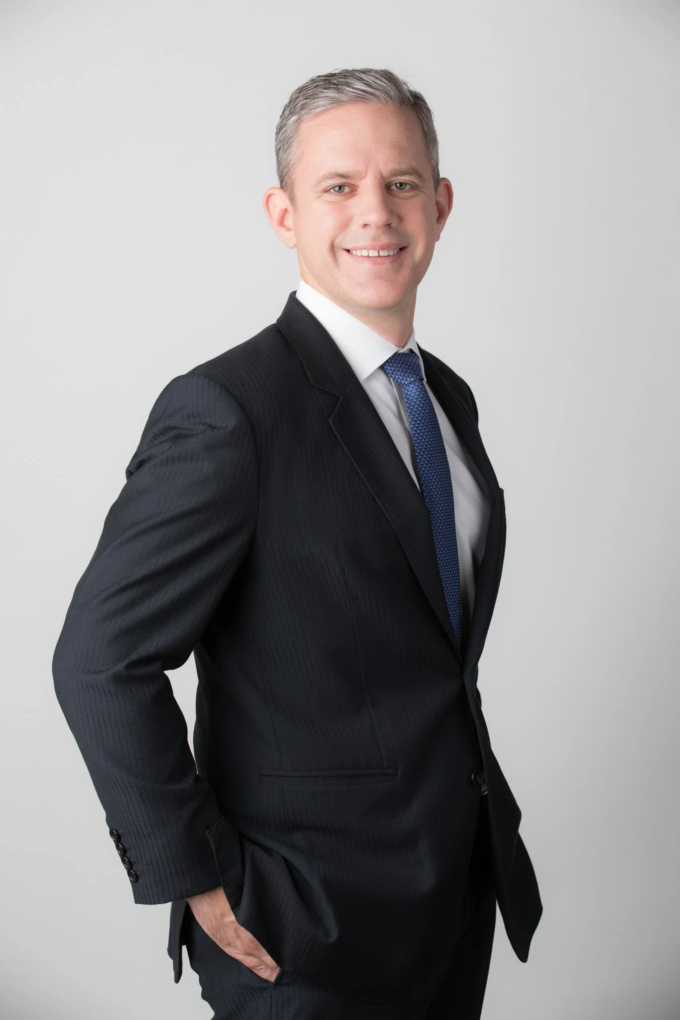 Executive portrait of a man in a black suit, white shirt, and blue tie standing against a plain light grey background.