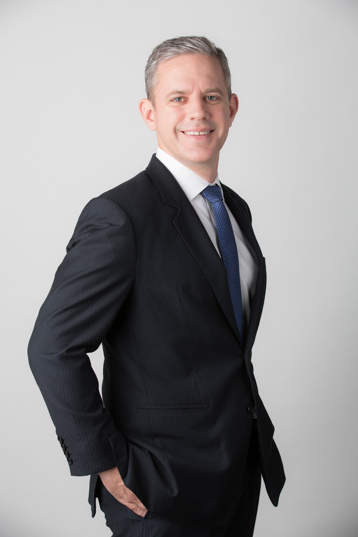 A man in a black suit, white shirt, and blue tie standing against a plain light grey background.