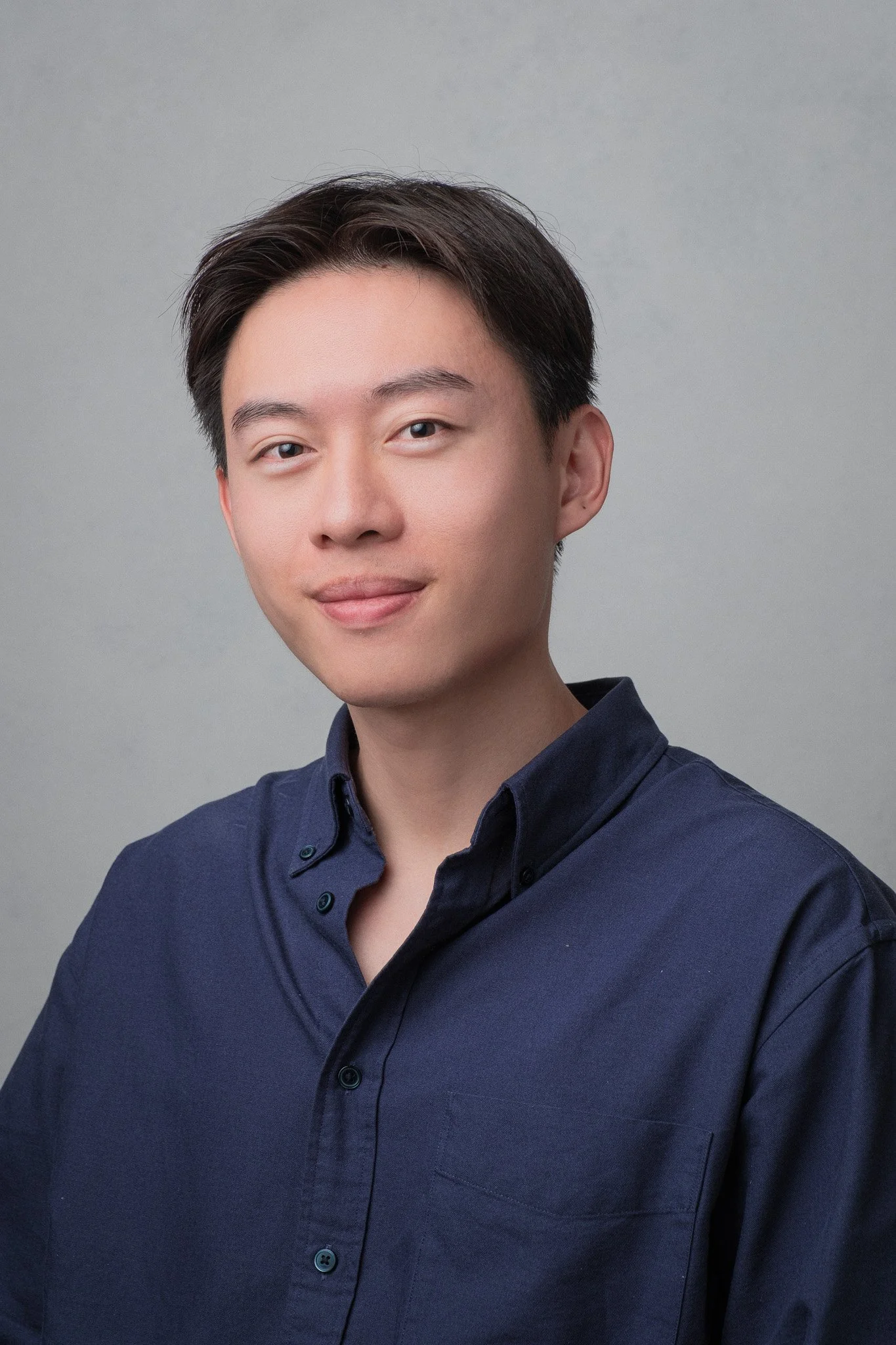 A young man with short dark hair, wearing a navy blue button-up shirt, is smiling gently and looking slightly to the right against a plain gray background.