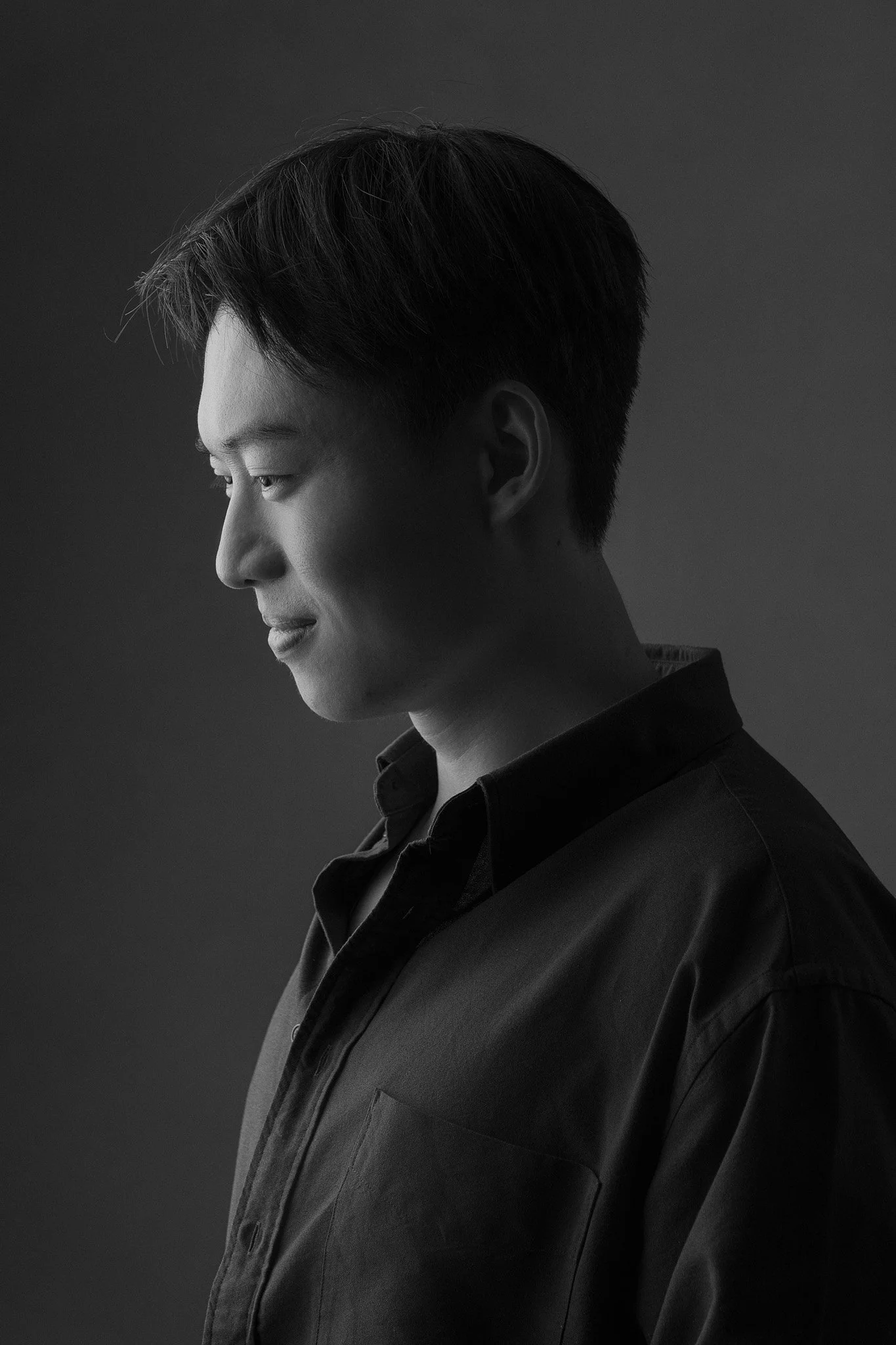 A black-and-white photo of a young man with short, styled hair, wearing a dark shirt, with a calm, contemplative expression, against a plain background.