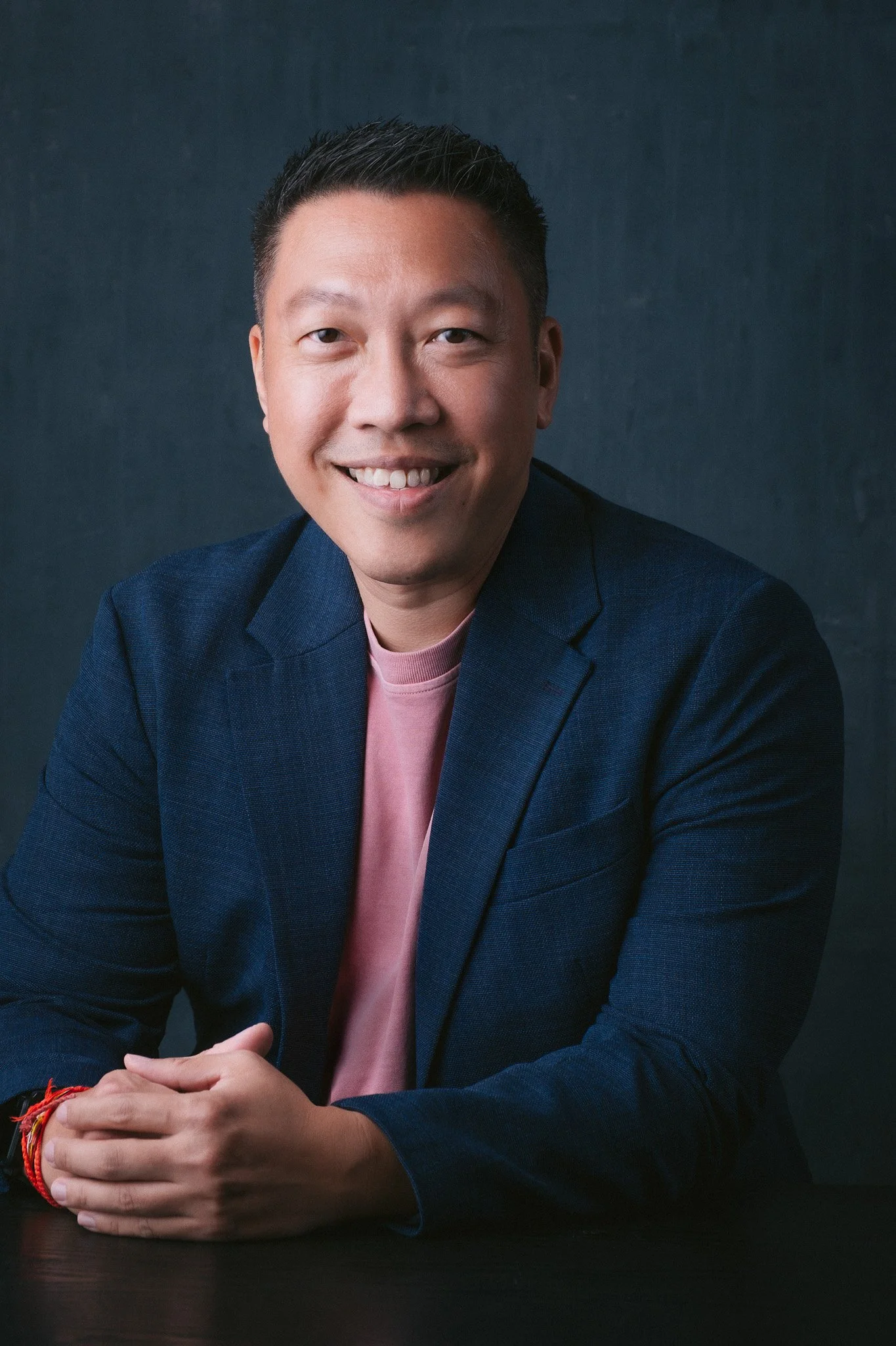 A man with short black hair, wearing a navy blazer and pink shirt, smiling and sitting at a black table against a dark background.