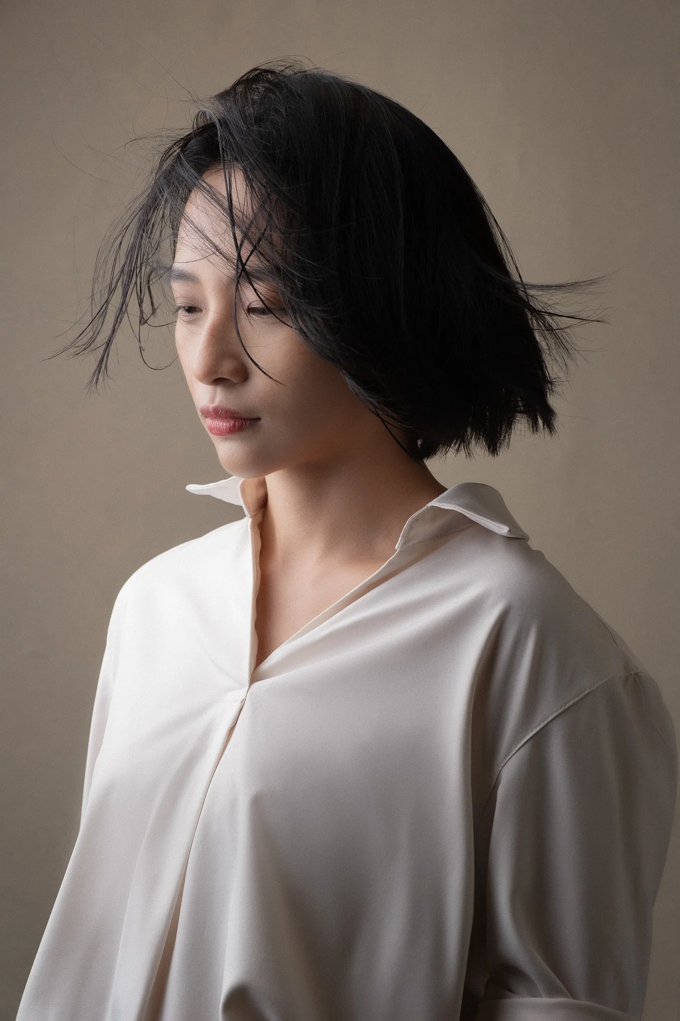 A woman with medium-length black hair and light skin wearing a white shirt with a collar, looking down with a neutral expression.