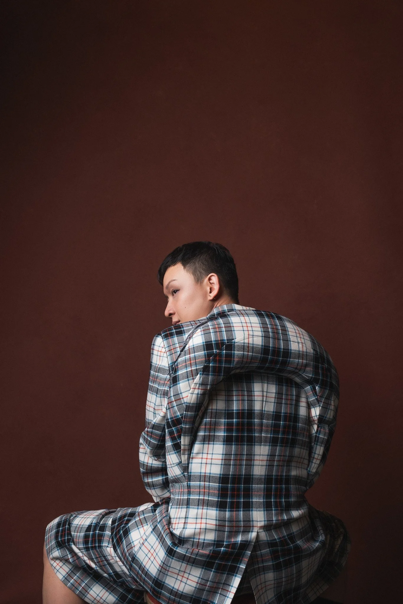Young person with short dark hair and pierced ears wearing a plaid shirt, seated on a leather chair against a plain brown background.