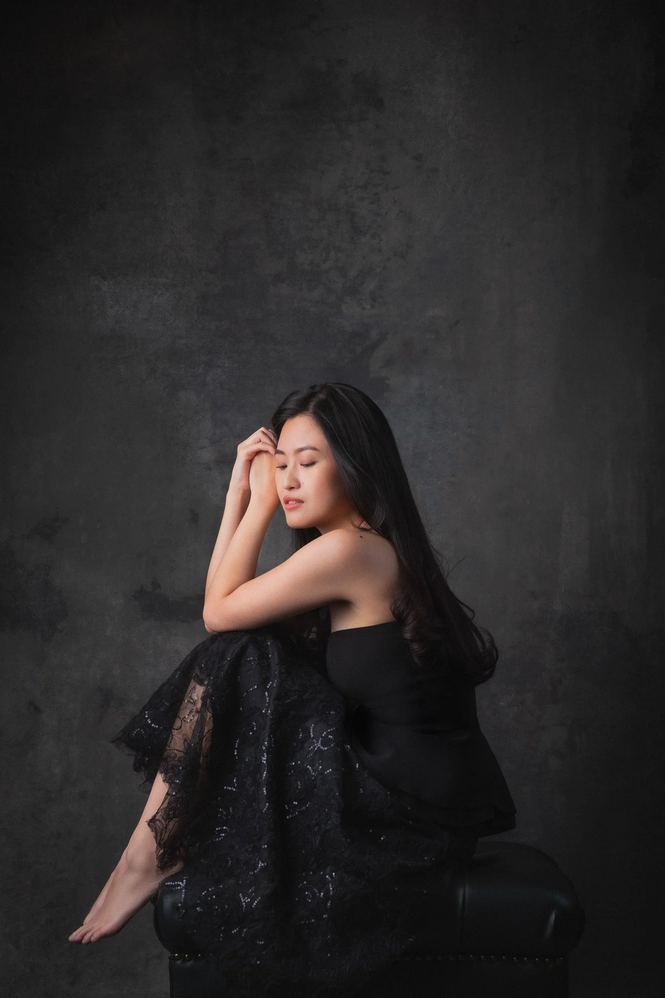 A woman with long black hair, wearing a black dress with lace and sequins, is sitting on a black stool against a dark textured background. She is posing with her eyes closed, head slightly tilted, and one hand resting on her forehead.
