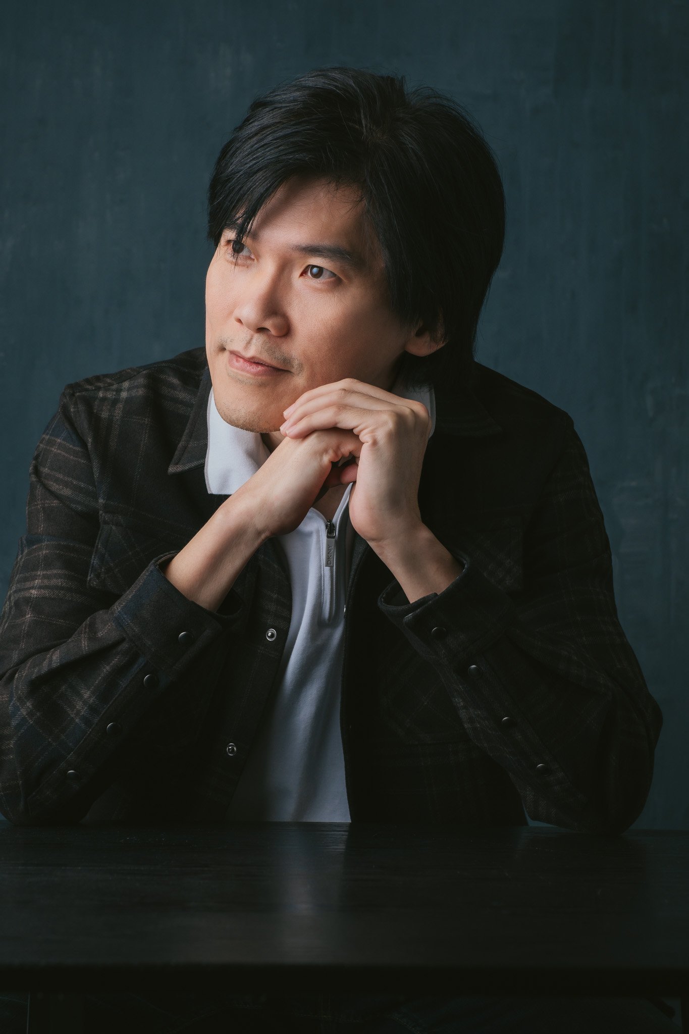 Close-up portrait of a man with black hair resting his chin on his hands against a dark background.