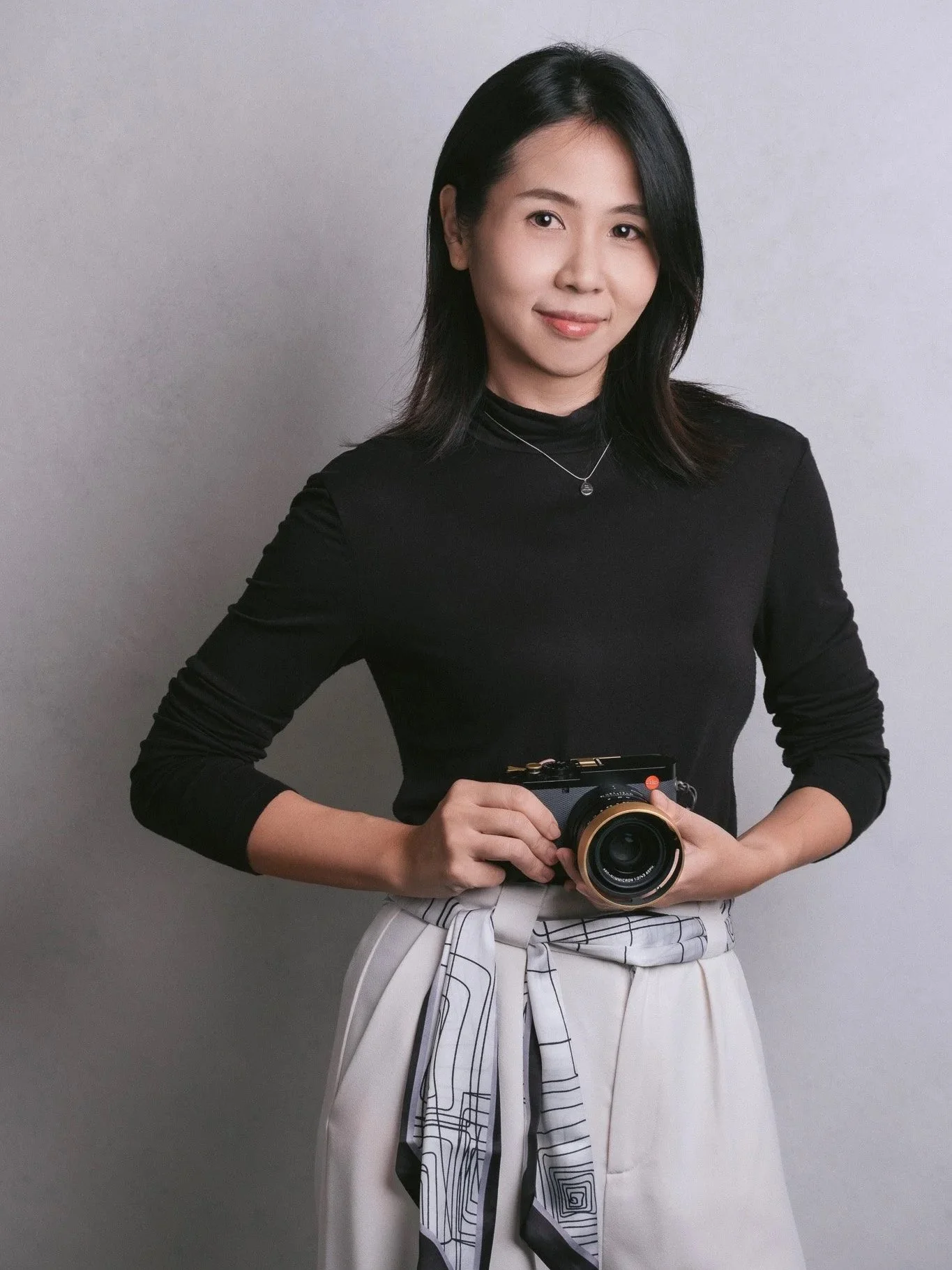 A woman with black hair wearing a black long-sleeve top, white patterned pants, and a plaid jacket tied around her waist, holding a camera in front of a plain gray background.