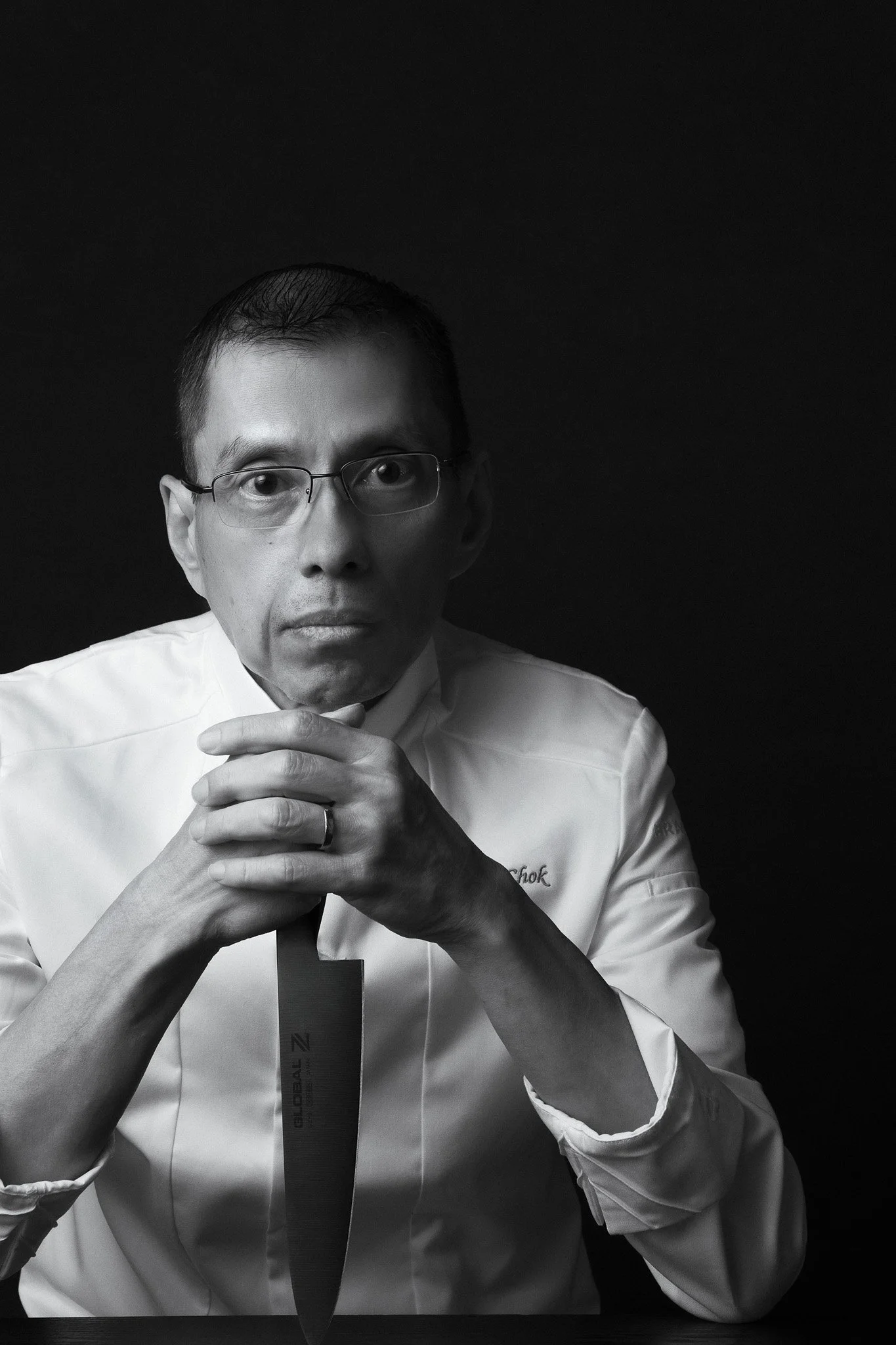 Black and white portrait of a man wearing glasses and a white chef's coat, holding a medal with a ribbon, against a dark background.