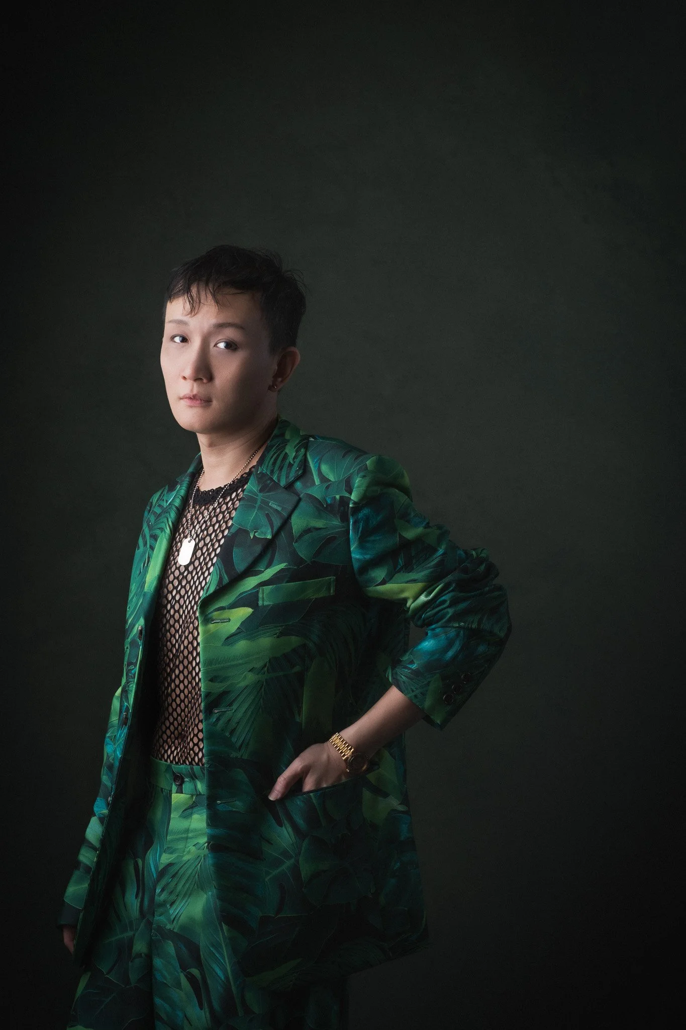 Person with short hair wearing a green tropical print suit over fishnet top, posing with one hand on hip against dark background.